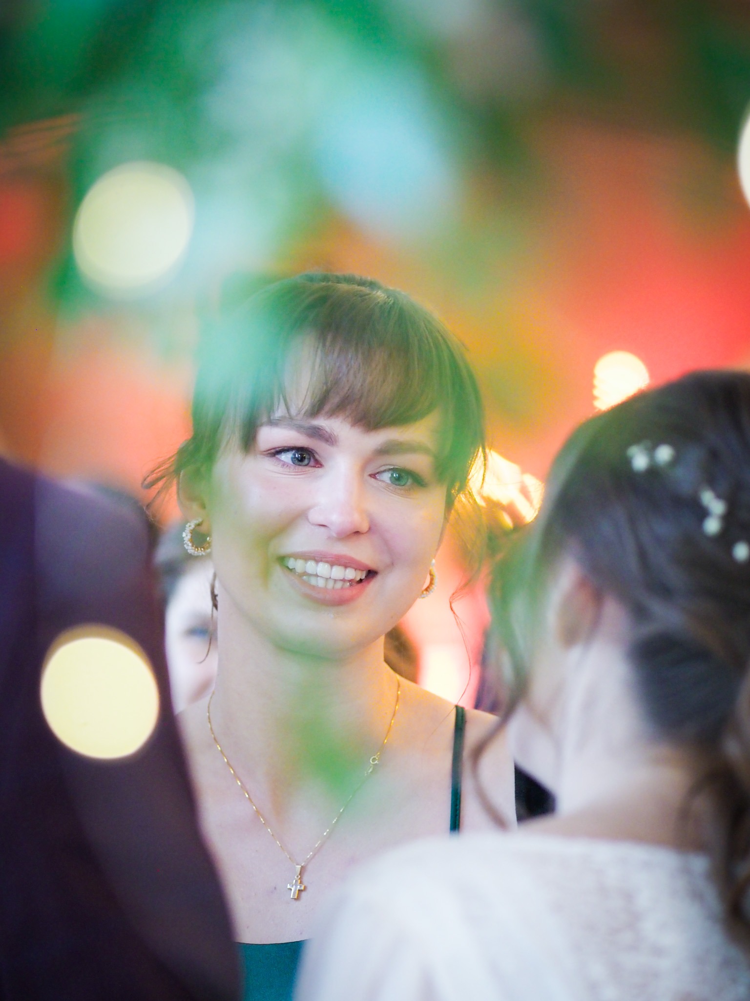 Uśmiechnięta kobieta z krótką grzywką, złotym naszyjnikiem z krzyżykiem i kolczykami, rozmawia z inną osobą podczas wydarzenia oświetlonego kolorowymi światłami.