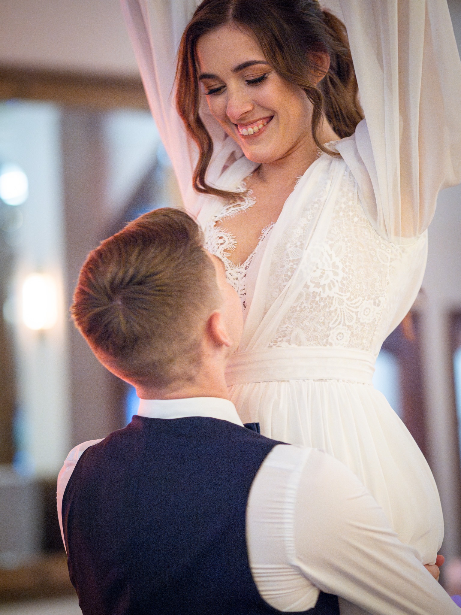 Uśmiechnięta panna młoda w koronkowej sukni ślubnej, podnoszona przez pana młodego w granatowej kamizelce podczas pierwszego tańca.