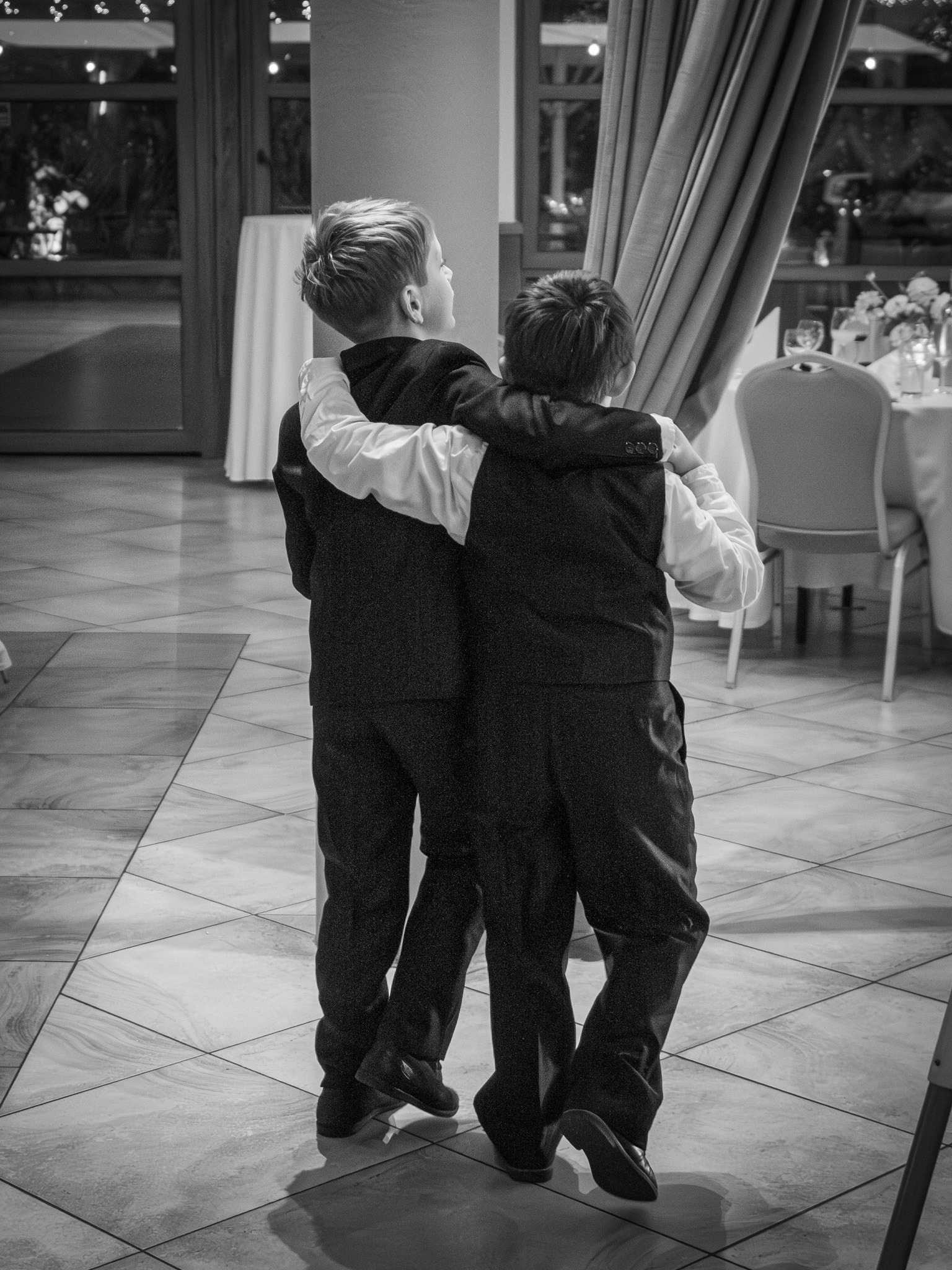 Czarno-białe zdjęcie dwóch chłopców w garniturach obejmujących się na parkiecie sali balowej, widok z tyłu, z widocznymi nakrytymi stołami i oknami w tle.