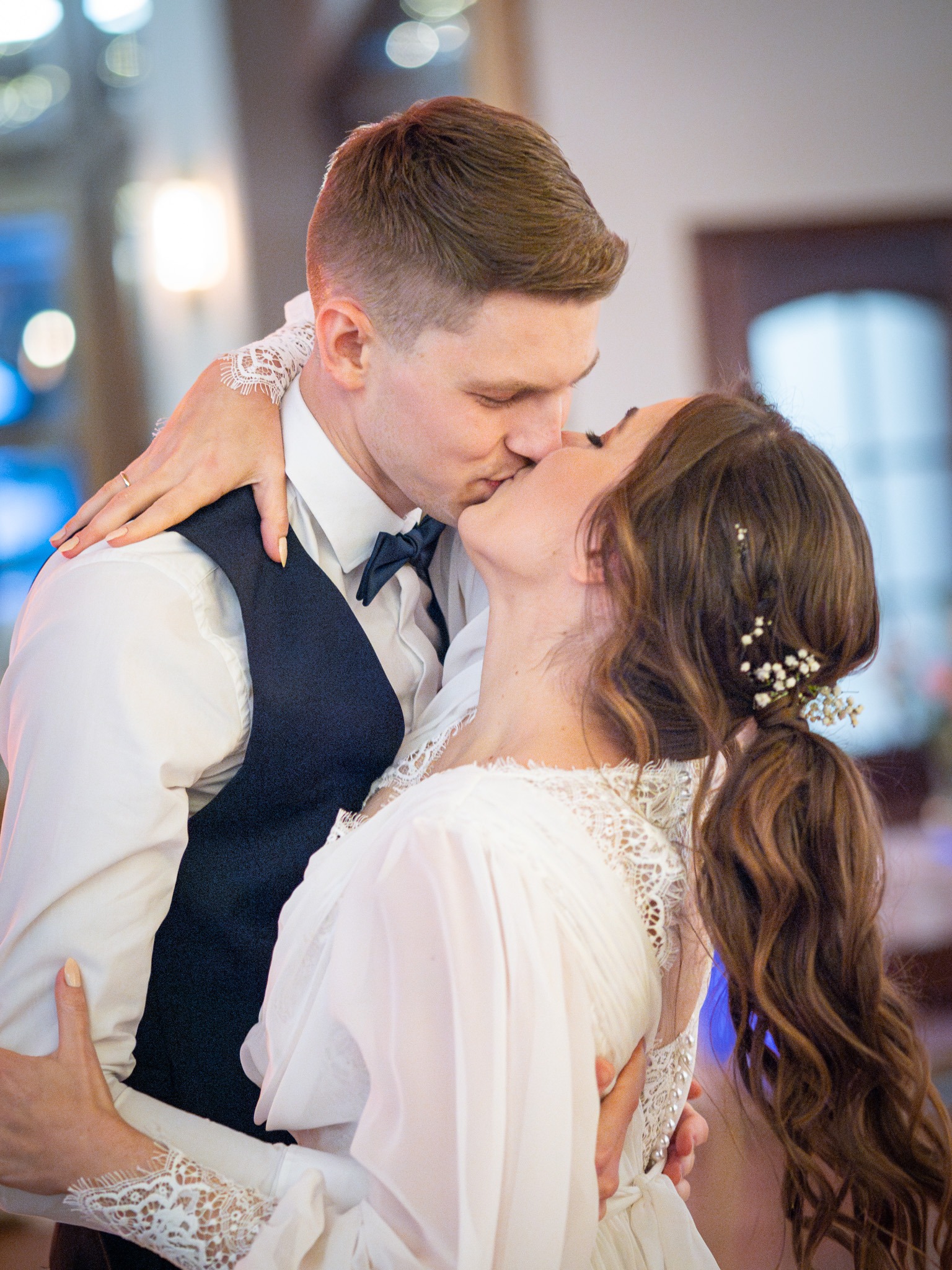 Pocałunek pary młodej: mężczyzna w czarnej kamizelce i muszce, kobieta w białej sukni z koronkowymi rękawami i kwiatami we włosach, w tle rozmyte światła.