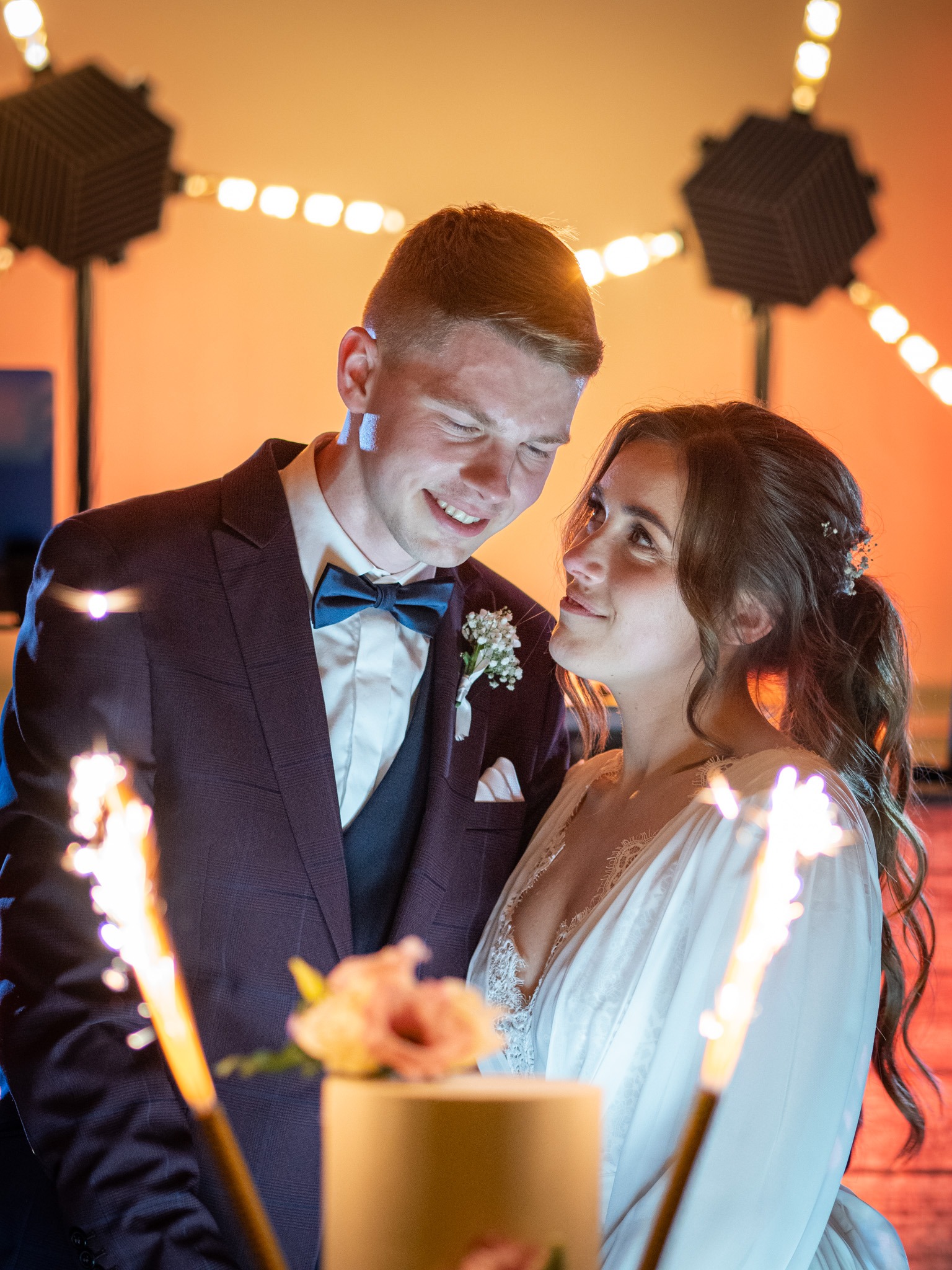 Uśmiechnięta para młoda, panna młoda patrzy na pana młodego, tort weselny z kwiatami i fontannami iskier, ciepłe oświetlenie sceniczne w tle.