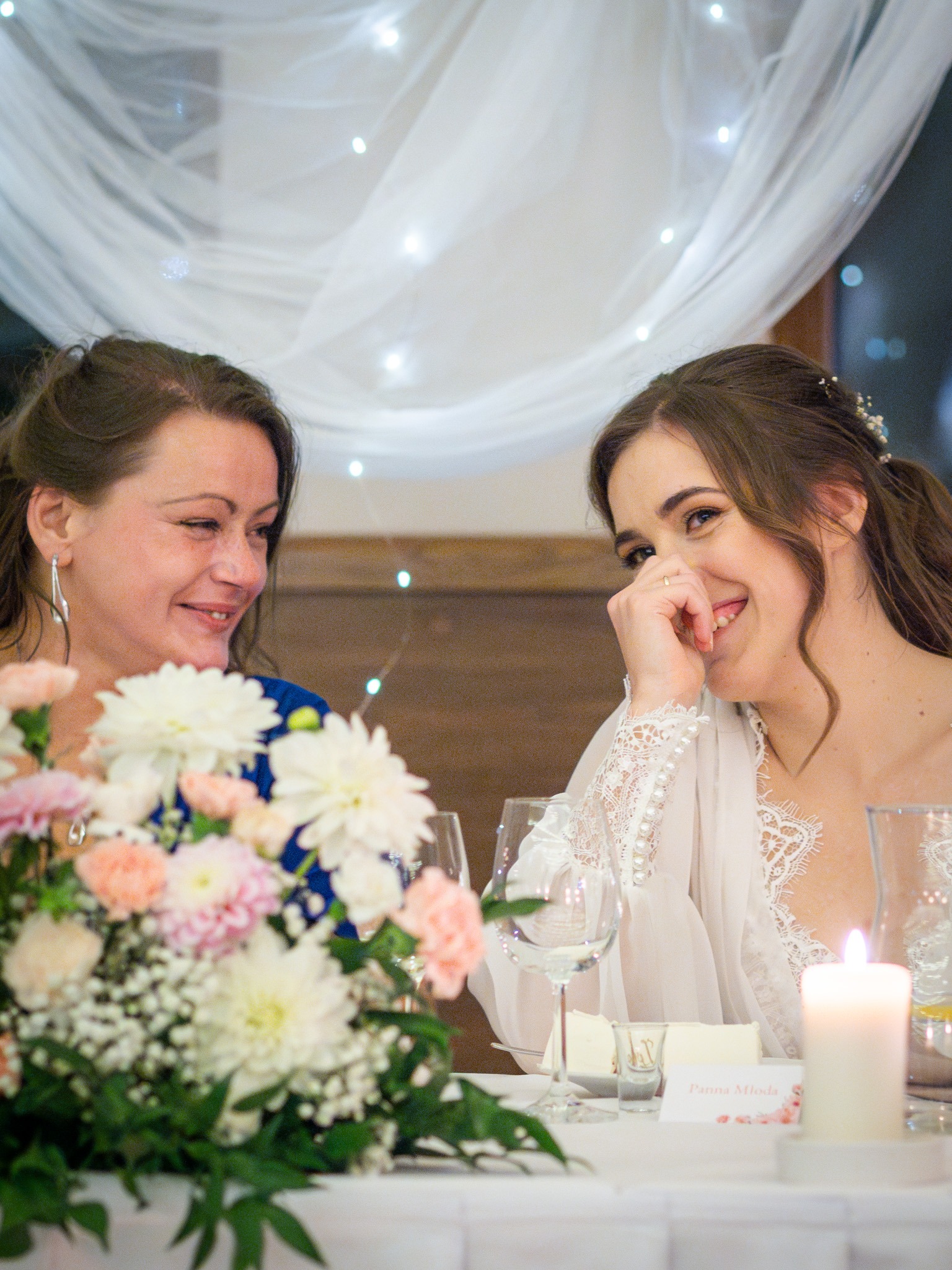 Uśmiechnięta panna młoda w białym szlafroku z koronką, siedząca przy stole z mamą, kwiatami, świecą i dekoracjami świetlnymi w tle.