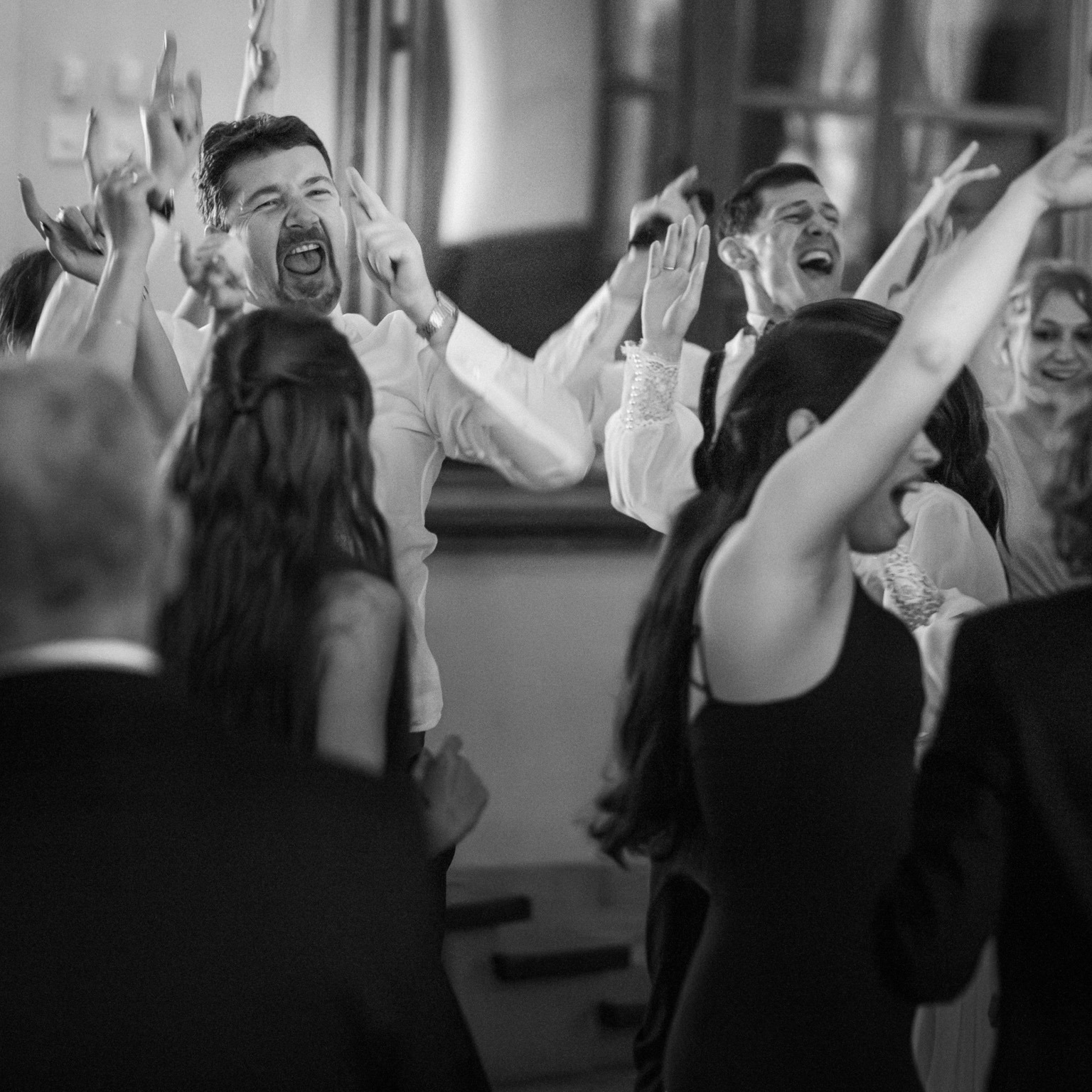 Czarno-białe zdjęcie rozbawionych gości weselnych podczas tańca, z uniesionymi rękami i radosnymi minami, uchwycone w dynamicznym ujęciu z bliska.