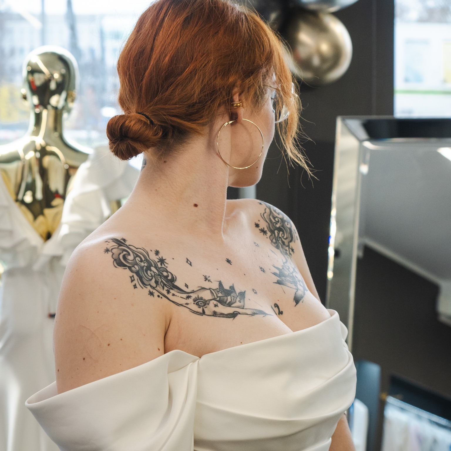 Kobieta z rudymi włosami upiętymi w kok, odwrócona tyłem, ubrana w białą suknię z odsłoniętymi ramionami. Widoczne tatuaże na dekolcie i ramieniu, złote kolczyki koła. W tle manekin i dekoracje.