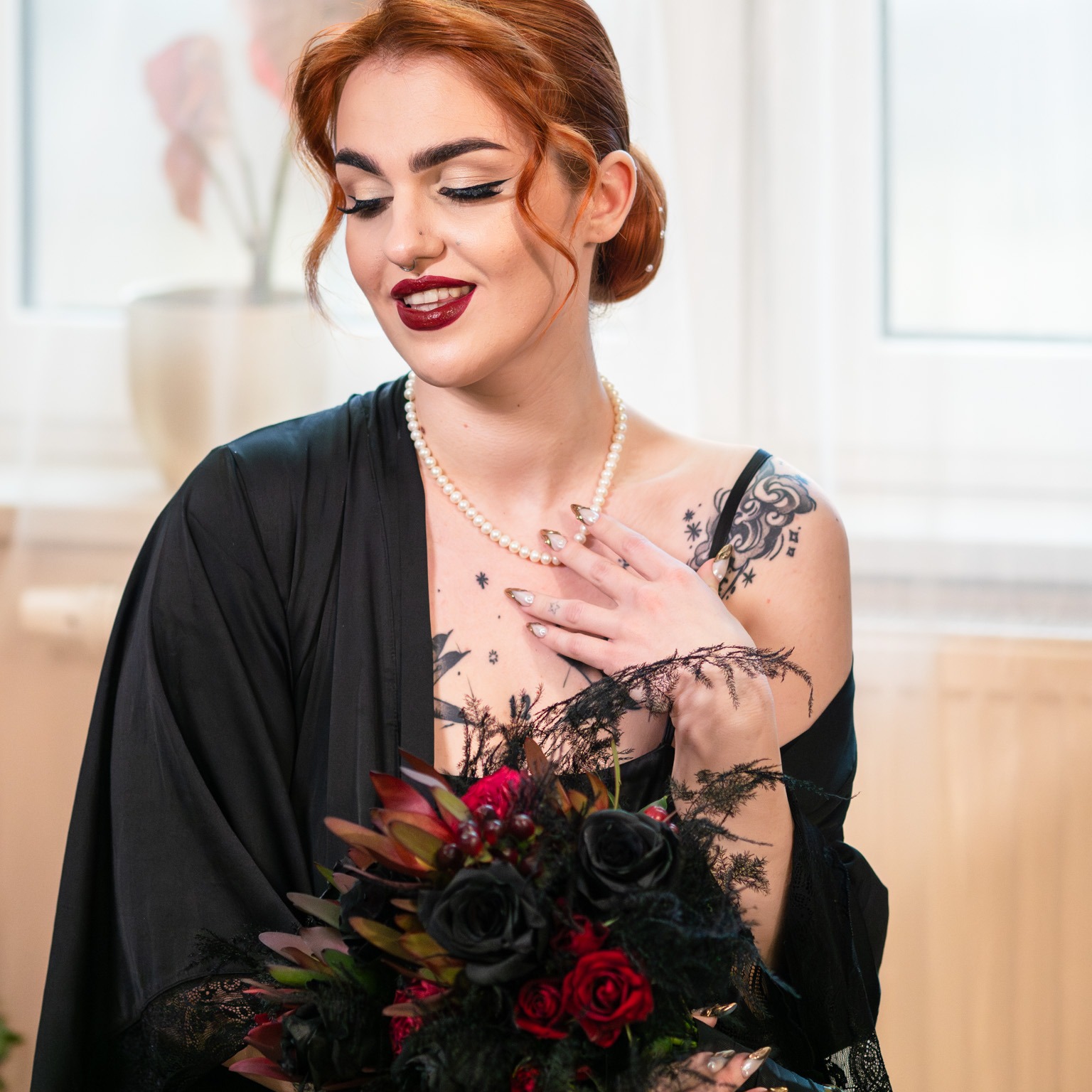 Kobieta z rudymi włosami, w czarnym szlafroku i perłowym naszyjniku, trzyma bukiet czarnych i czerwonych róż, z tatuażami na ramionach i dekolcie, patrzy w dół z delikatnym uśmiechem.