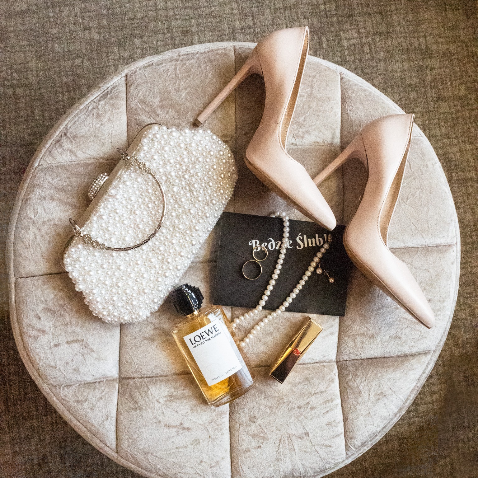 Przygotowania ślubne: beżowe szpilki, perłowa torebka, perfumy Loewe, szminka, biżuteria i zaproszenie na pufie w odcieniach beżu.