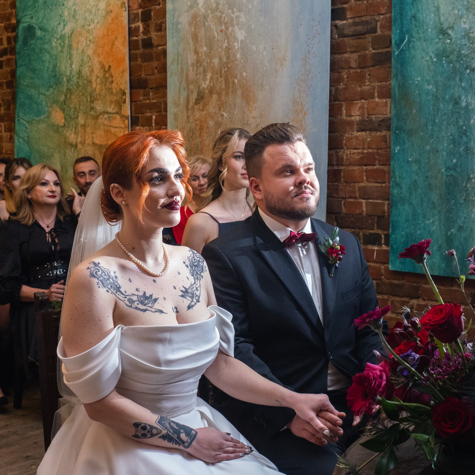 Para młoda siedzi obok siebie podczas ceremonii, panna młoda z rudymi włosami i tatuażami na dekolcie, pan młody w czarnym smokingu z bordową muchą, w tle ceglana ściana i abstrakcyjne obrazy.