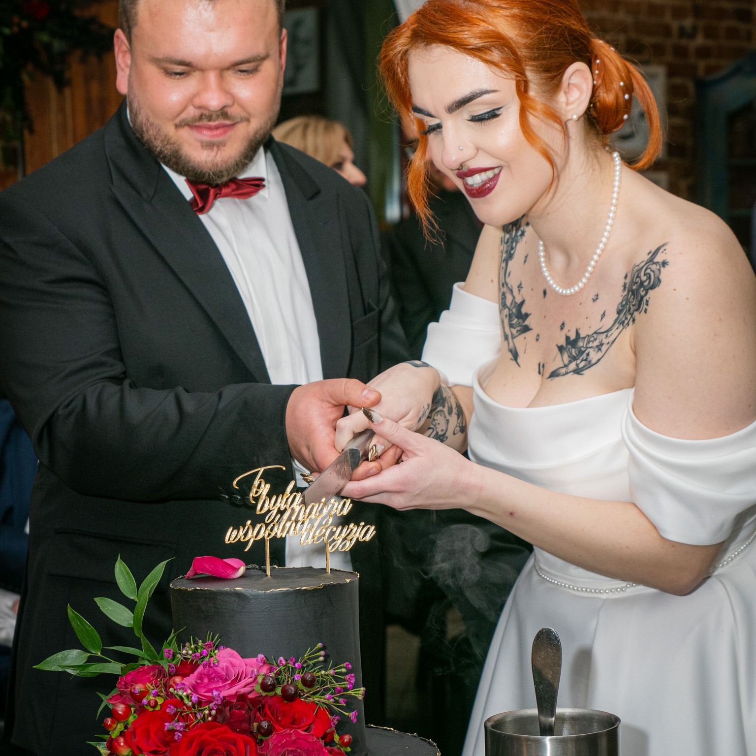 Para młoda kroi czarny tort weselny z napisem 'To była najpiękniejsza wspólna decyzja', zdobiony czerwonymi różami i zielonymi liśćmi. Panna młoda ma tatuaże i perły, pan młody w czarnym smokingu...