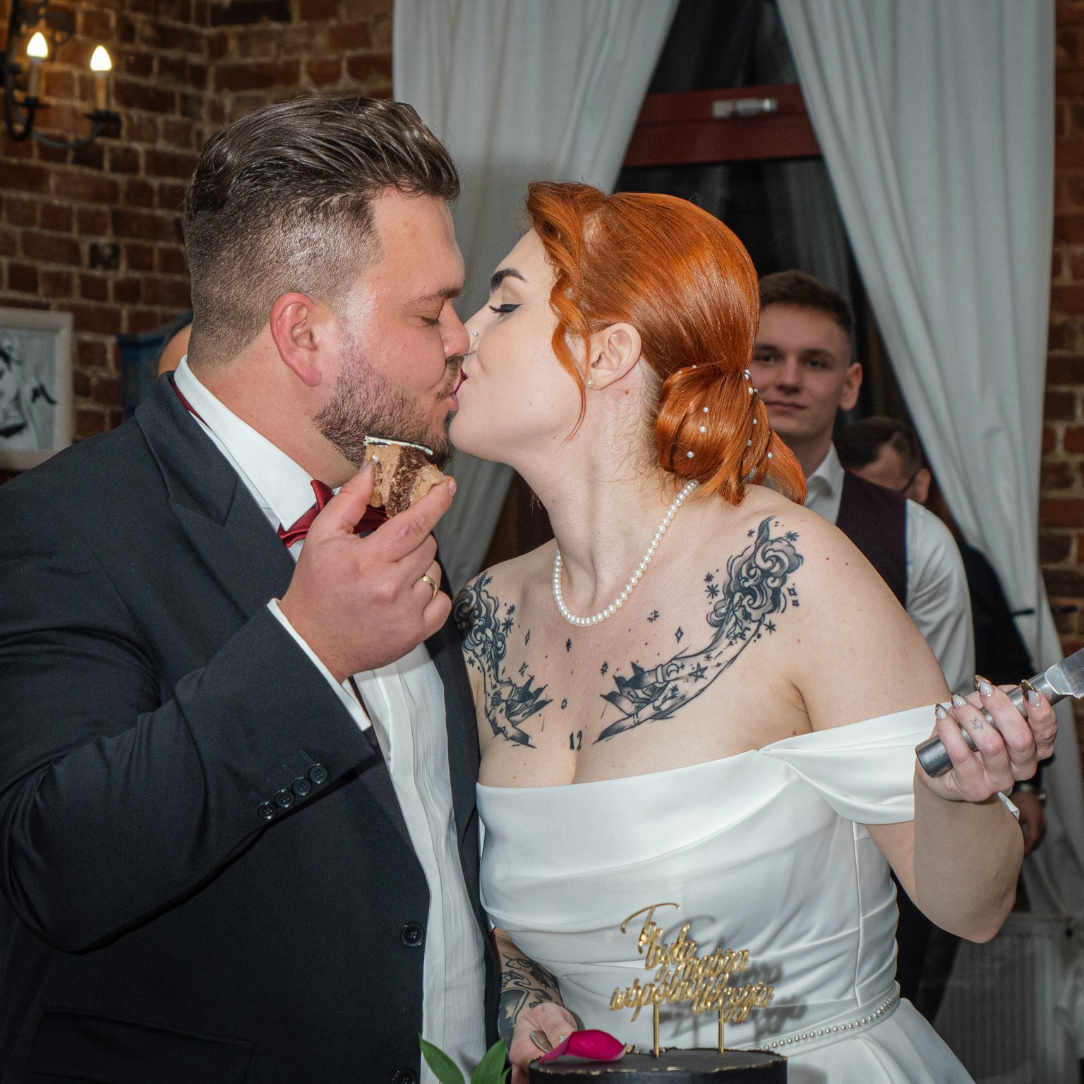 Para młoda całuje się, jedząc kawałek tortu weselnego. Panna młoda ma rude włosy upięte w kok, białą suknię i tatuaże na dekolcie. Pan młody w czarnym garniturze i bordowej muszce. W tle ceglana...
