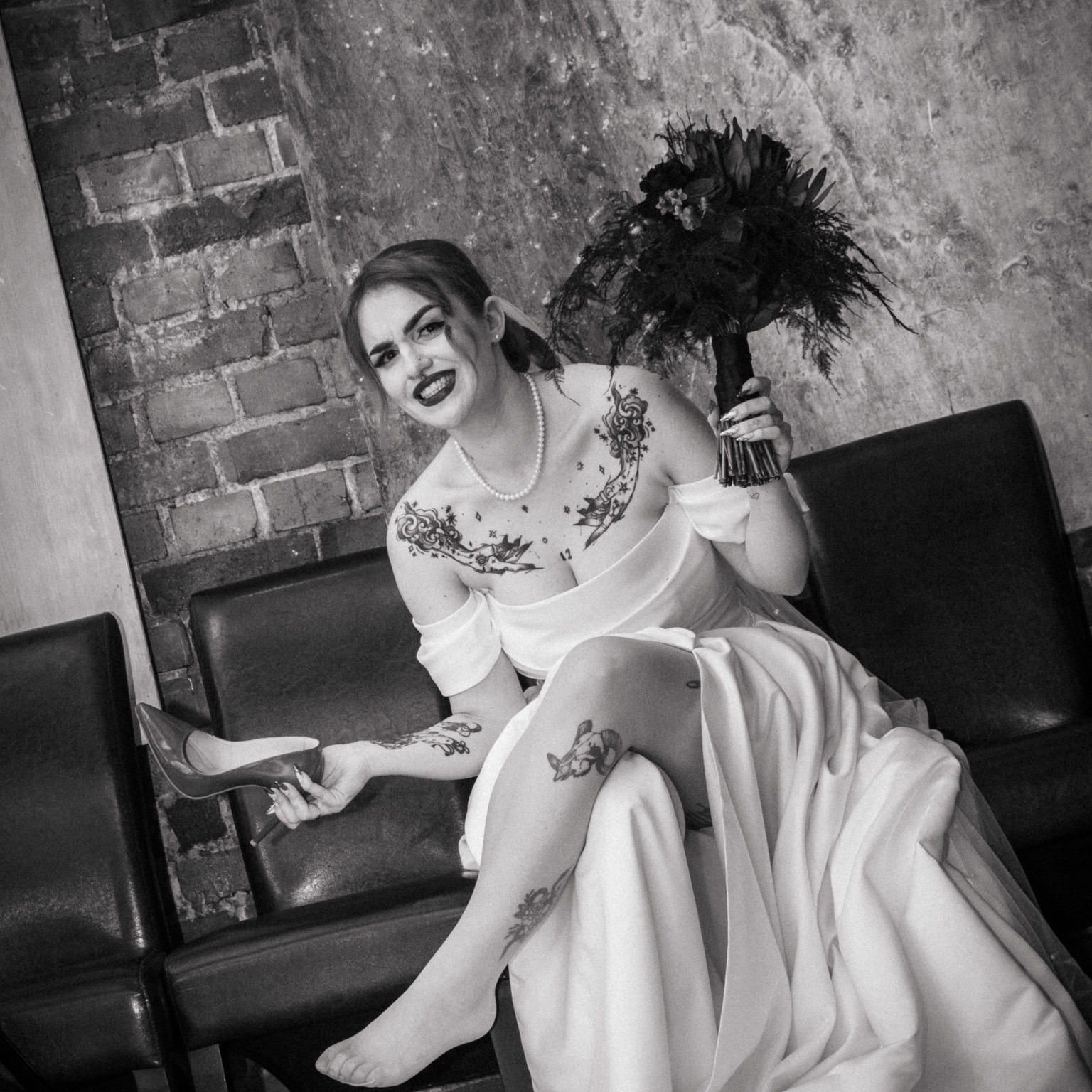 Czarno-białe zdjęcie kobiety w sukni ślubnej, siedzącej na krześle z bukietem w dłoni, trzymającej czerwony szpilkę. Widoczne tatuaże na ciele, ciemna szminka i perły na szyi. W tle ceglana ściana.
