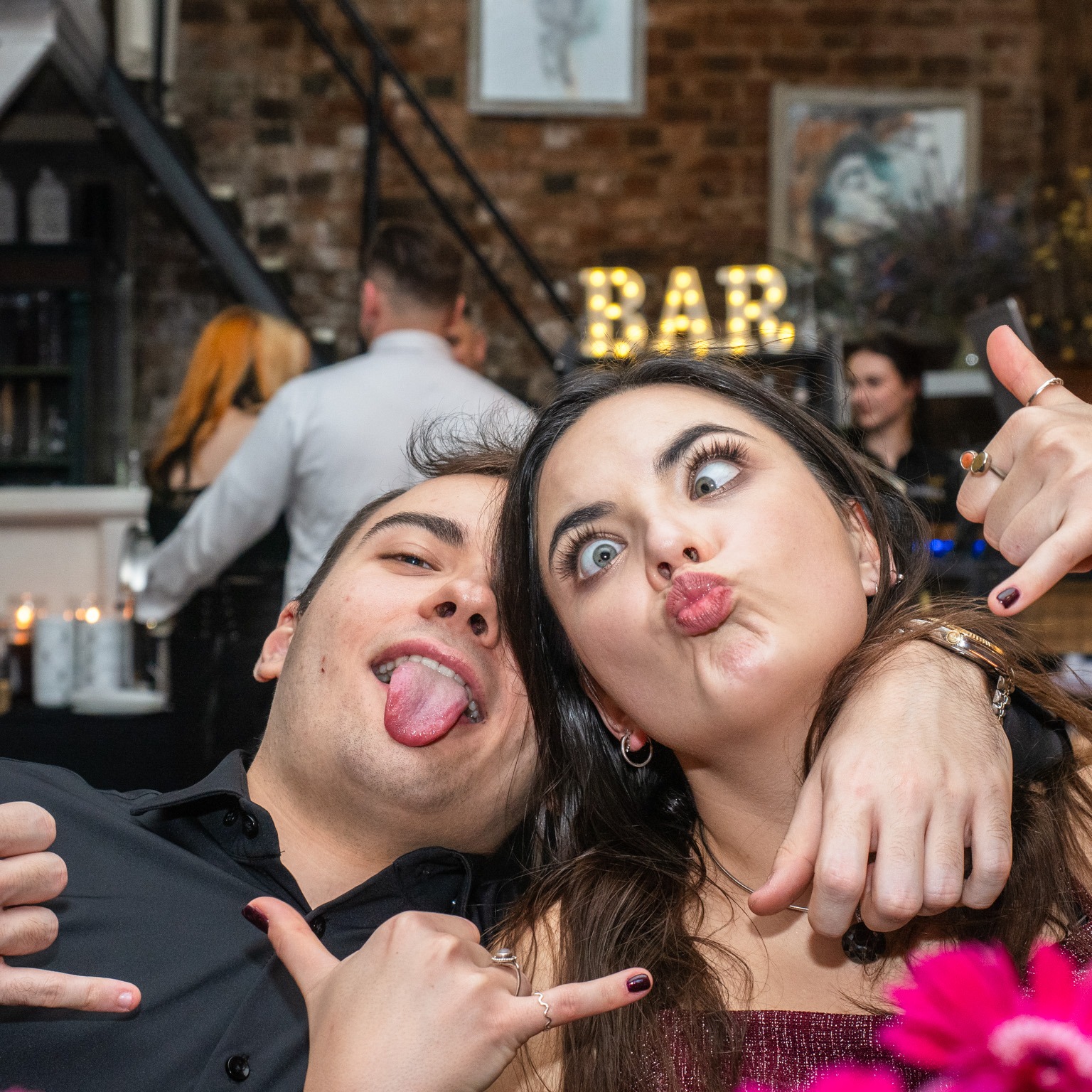 Para robiąca zabawne miny w barze, mężczyzna wystawia język, kobieta robi zeza i dzióbek, w tle napis 'BAR' z żarówek.