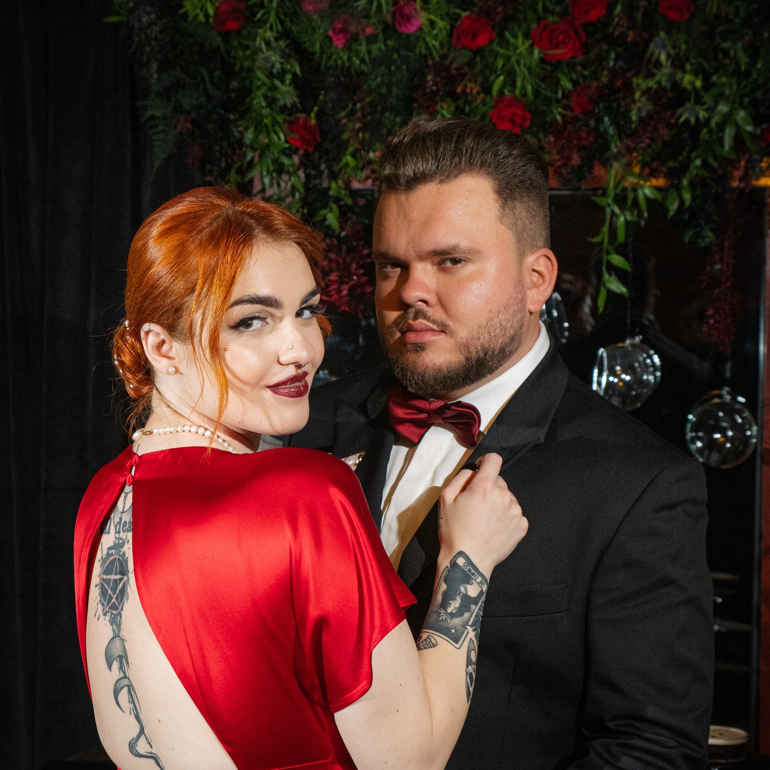 Elegancka para w formalnych strojach pozuje na tle dekoracji z czerwonych róż i błyszczących kul, kobieta w czerwonej sukni z tatuażami na plecach, mężczyzna w czarnym smokingu i bordowej muszce.