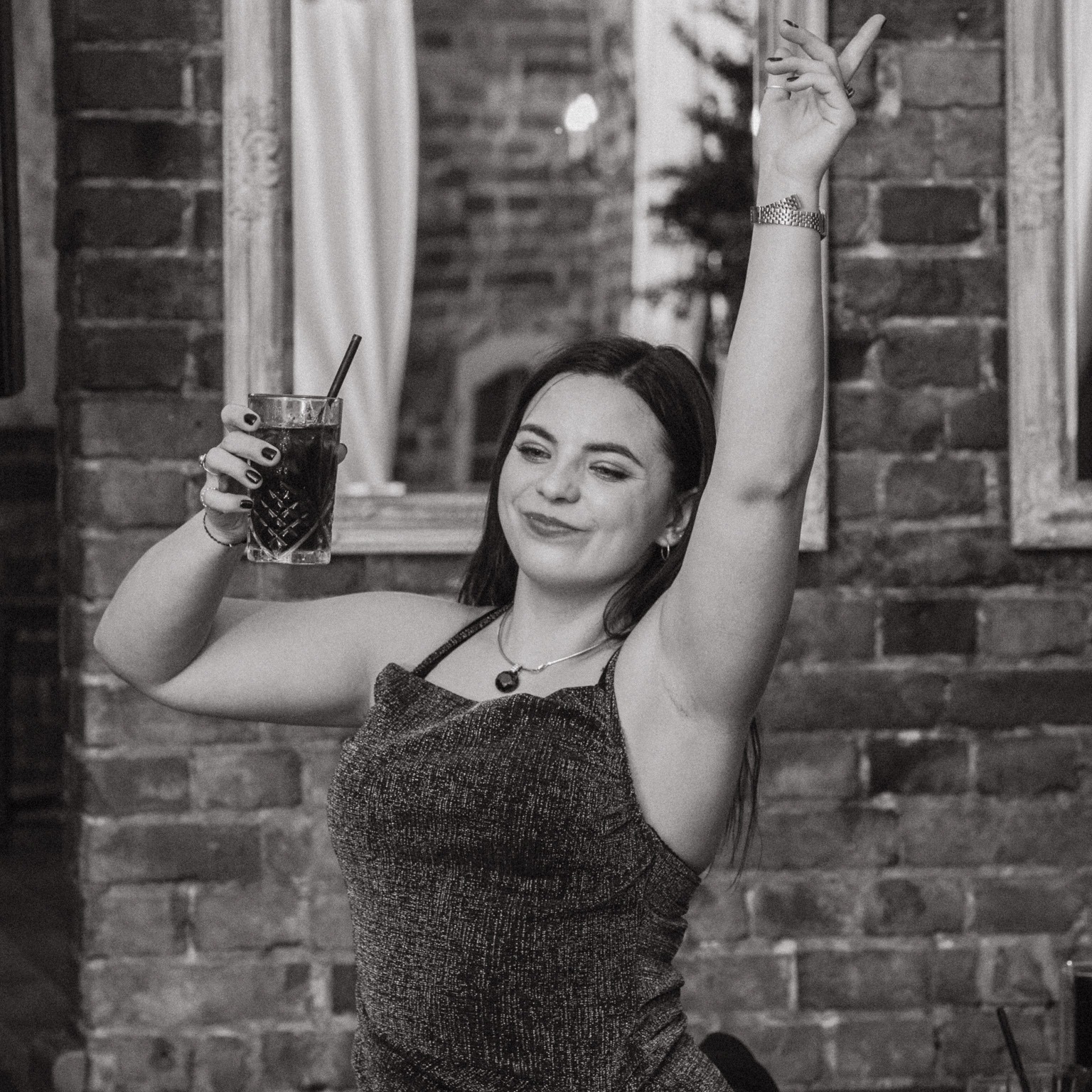 Kobieta z drinkiem w błyszczącej sukience, pozująca na tle ceglanej ściany we Wrocławiu, z uniesioną ręką i uśmiechem.