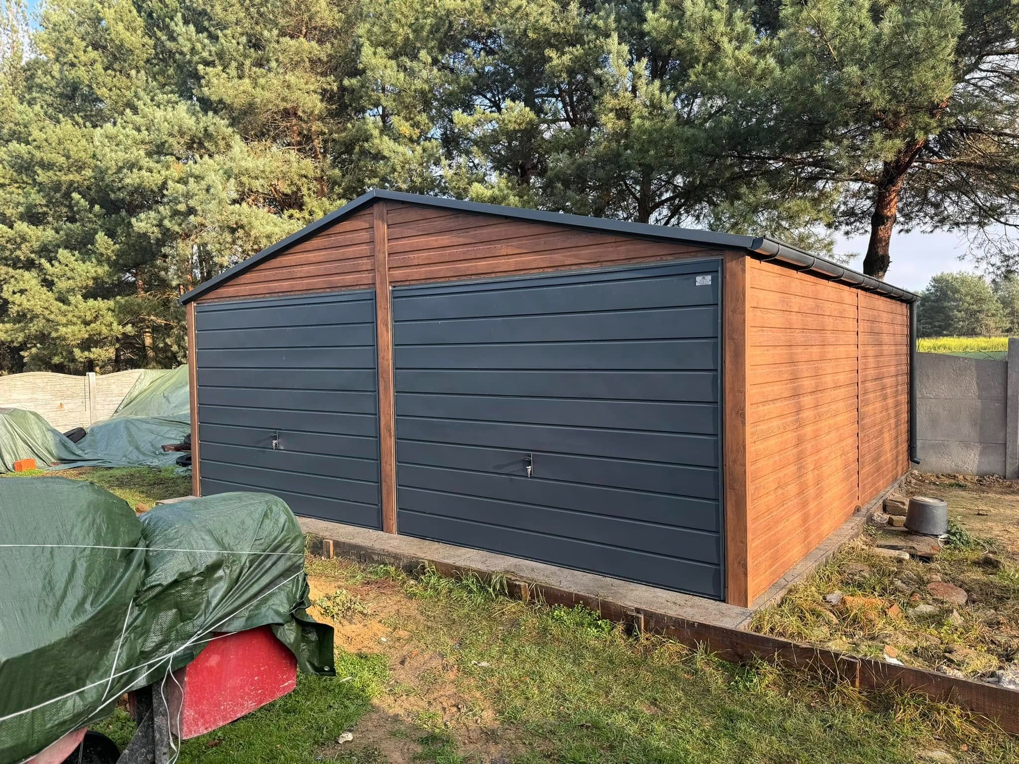 Dwustanowiskowy garaż z blachy trapezowej w kolorze grafitowym, z drewnopodobną elewacją, na tle iglastego lasu i betonowego ogrodzenia.