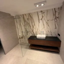 Nowoczesna łazienka z kabiną prysznicową typu walk-in, marmurową ścianą i drewnianą szafką pod umywalką. Minimalistyczny design i jasna kolorystyka.