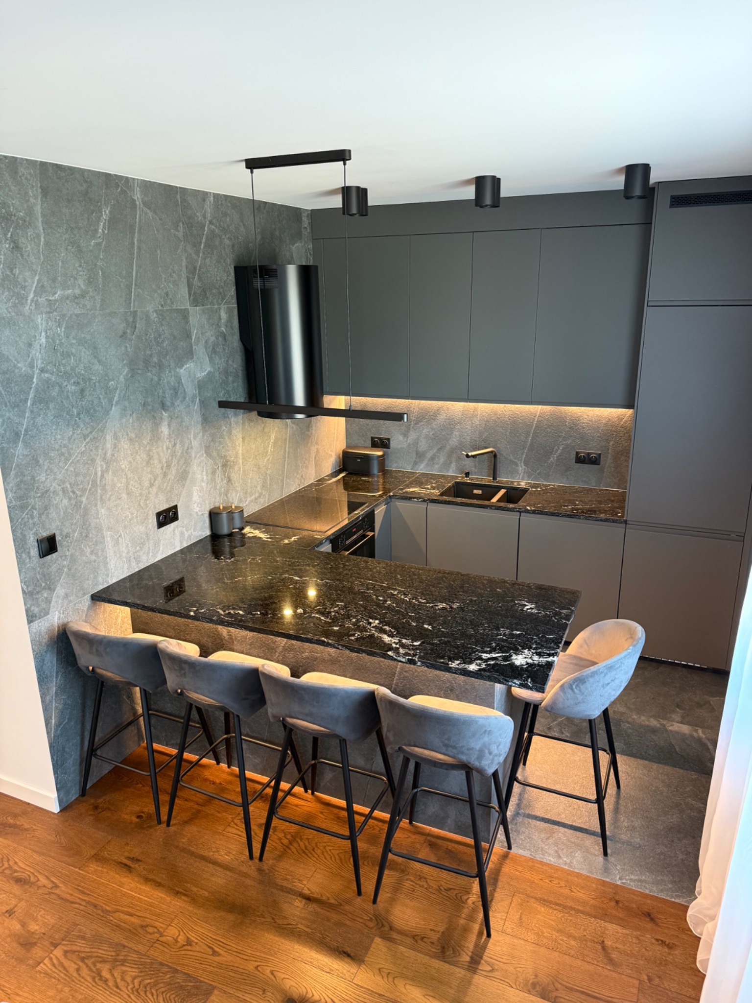 Nowoczesna kuchnia z szarymi szafkami, czarnym blatem i marmurową ścianą. Wysokie krzesła barowe z szarym obiciem. Oświetlenie punktowe i drewniana podłoga.