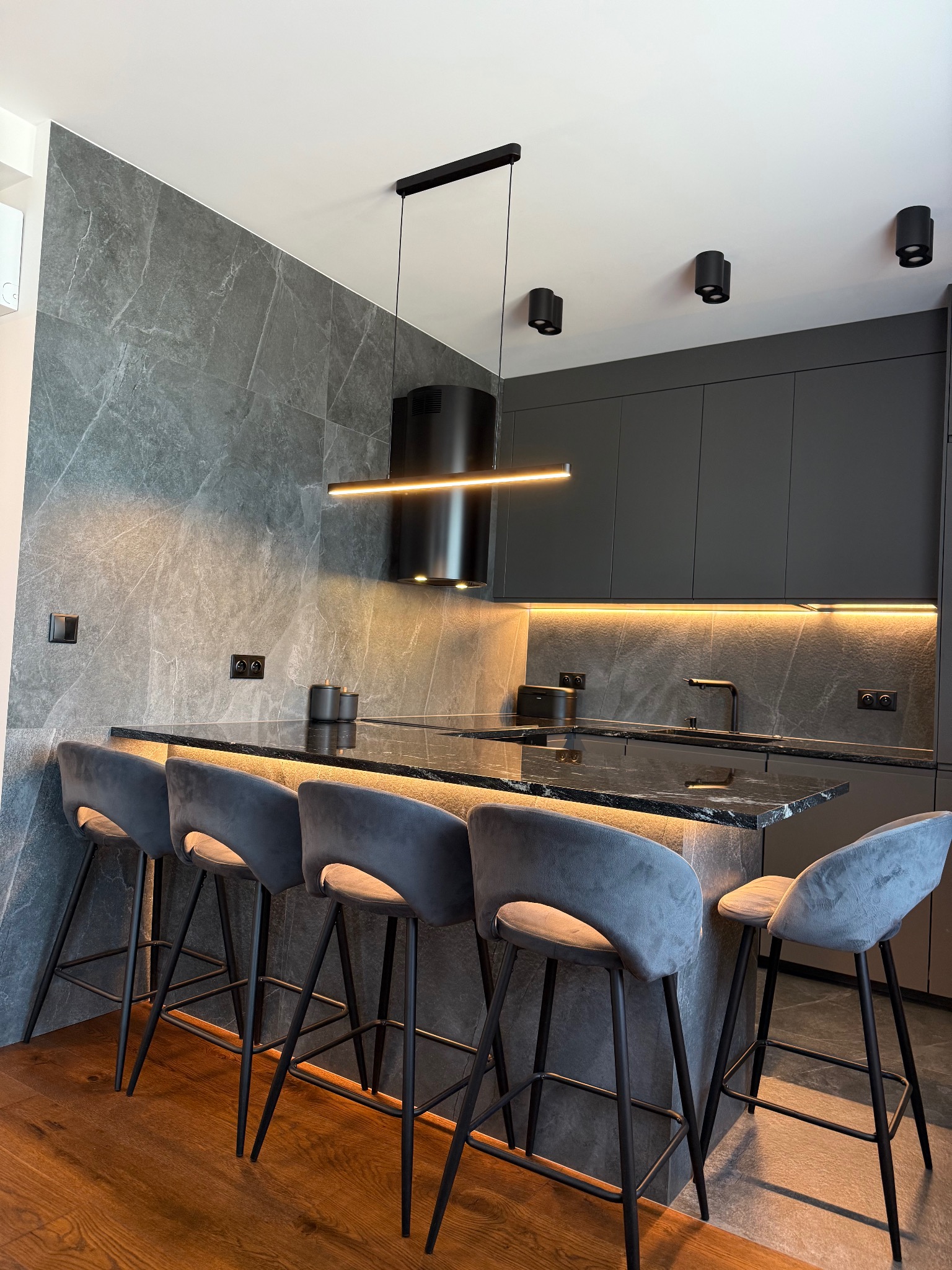 Nowoczesna kuchnia w odcieniach szarości z marmurową ścianą, oświetleniem LED i eleganckimi hokerami. Minimalistyczny design z czarnymi akcentami i drewnianą podłogą.