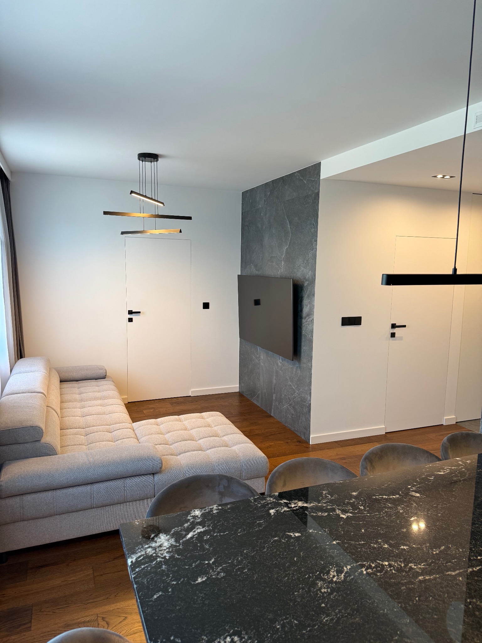 Nowoczesne wnętrze: szary narożnik, stół z czarnym blatem, ściana z szarego kamienia, telewizor, białe drzwi i designerska lampa wisząca.