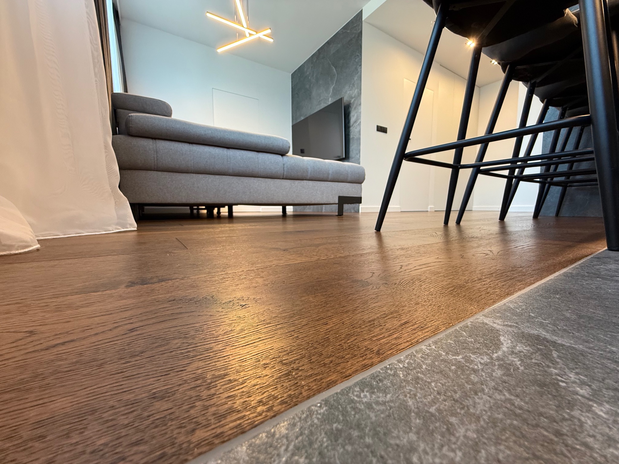 Nowoczesne wnętrze z drewnianą podłogą, szarą sofą i czarnymi krzesłami. Widok z niskiej perspektywy podkreśla fakturę podłogi i minimalistyczny design.