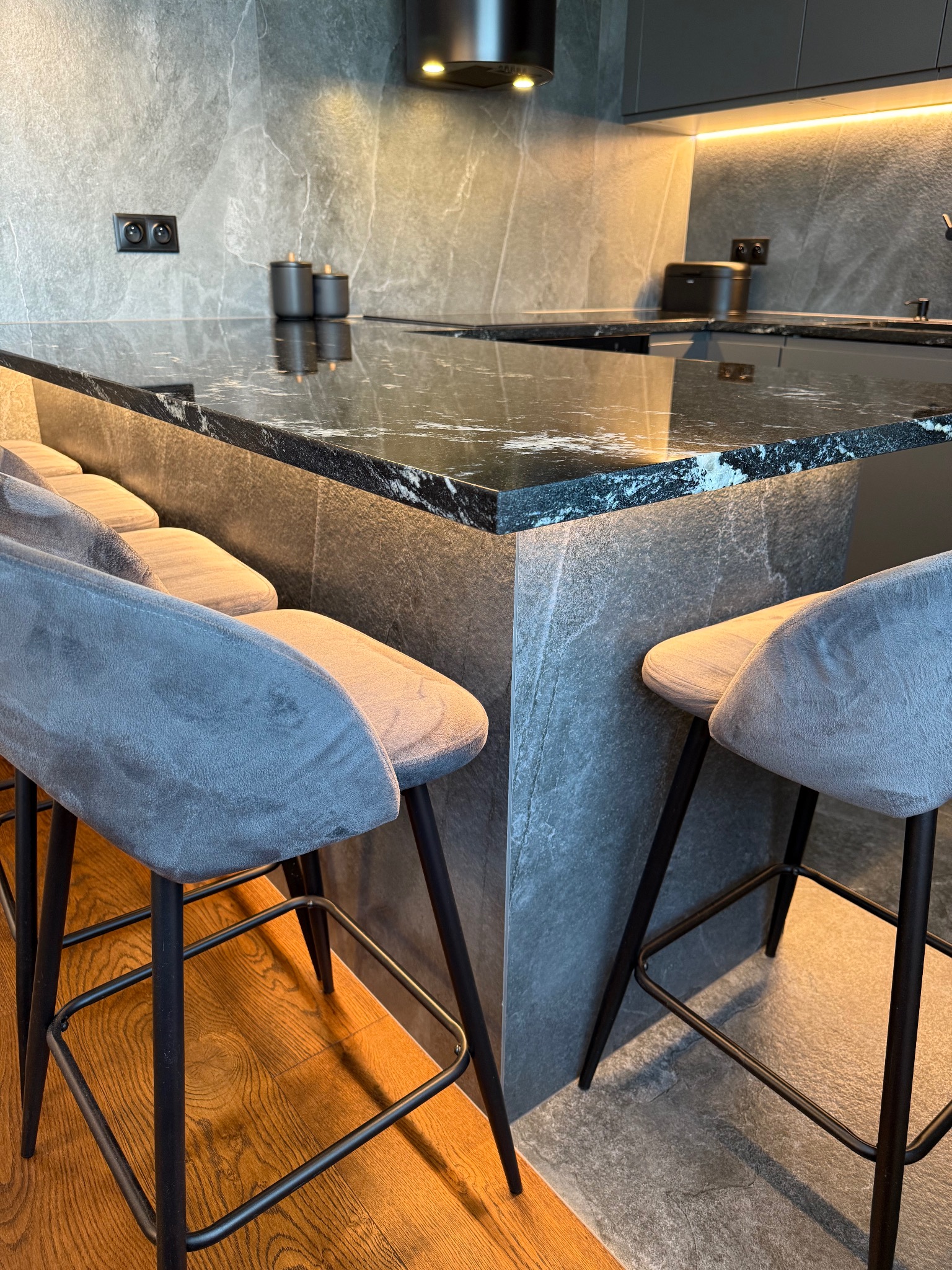 Nowoczesna kuchnia z wyspą i hokerami. Blat z ciemnego kamienia z wyraźnym użyleniem, szare ściany i meble. Oświetlenie LED pod szafkami dodaje ciepła.