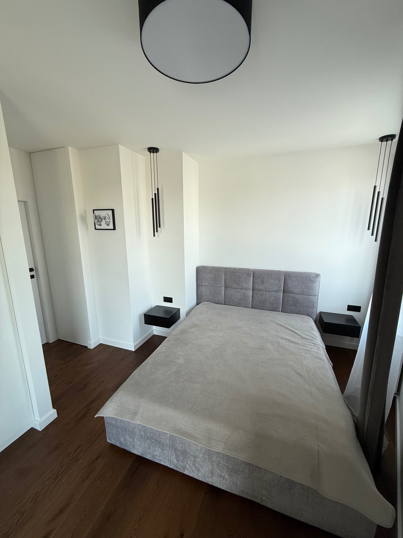 Nowoczesna sypialnia z szarym łóżkiem, minimalistycznymi lampami wiszącymi i ciemną podłogą. Na ścianie obrazek w czarnej ramce i czarne szafki nocne.