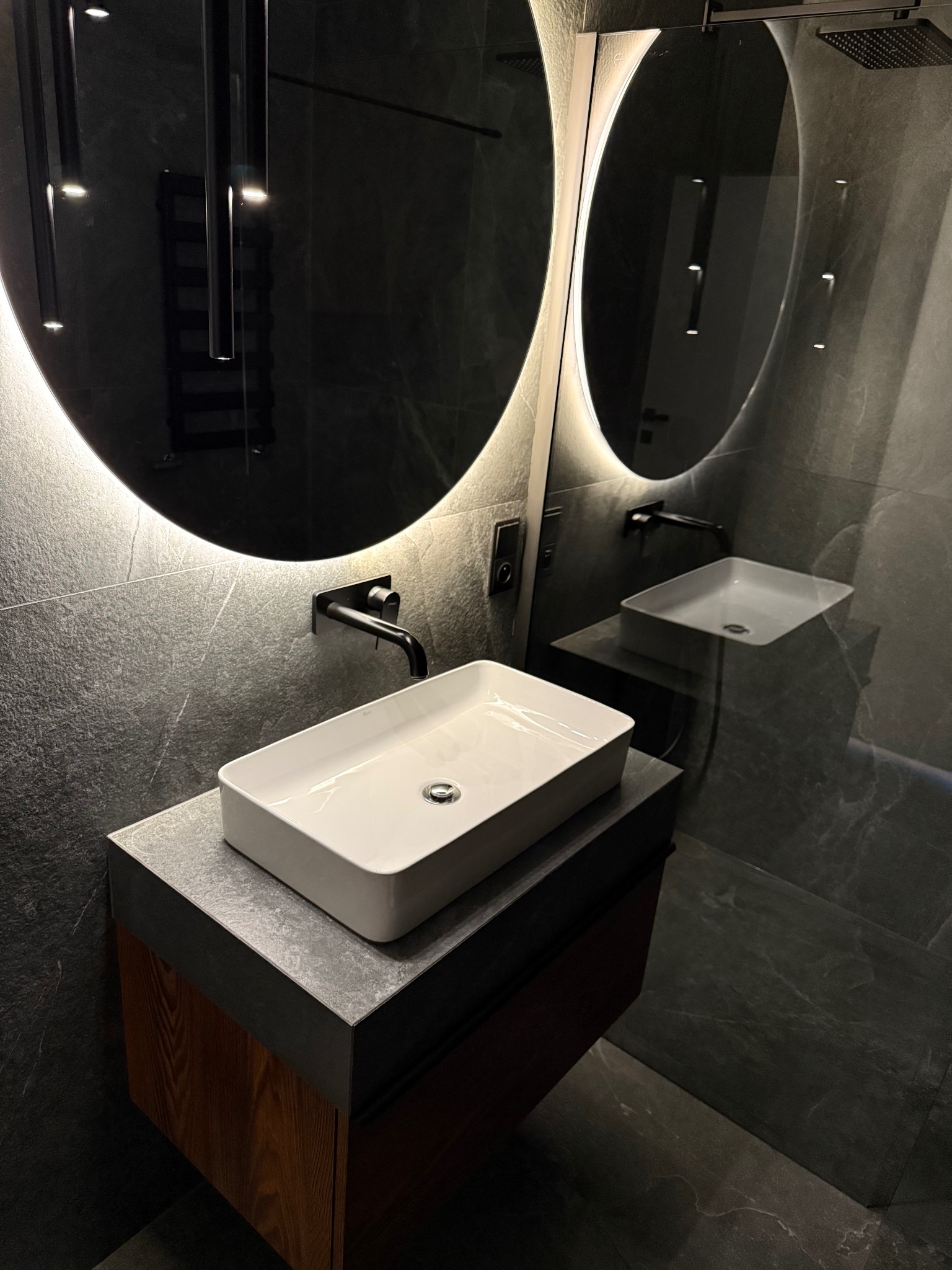Nowoczesna łazienka w ciemnej tonacji z podświetlanym okrągłym lustrem, prostokątną umywalką nablatową i drewnianą szafką pod umywalką. Minimalistyczny design.