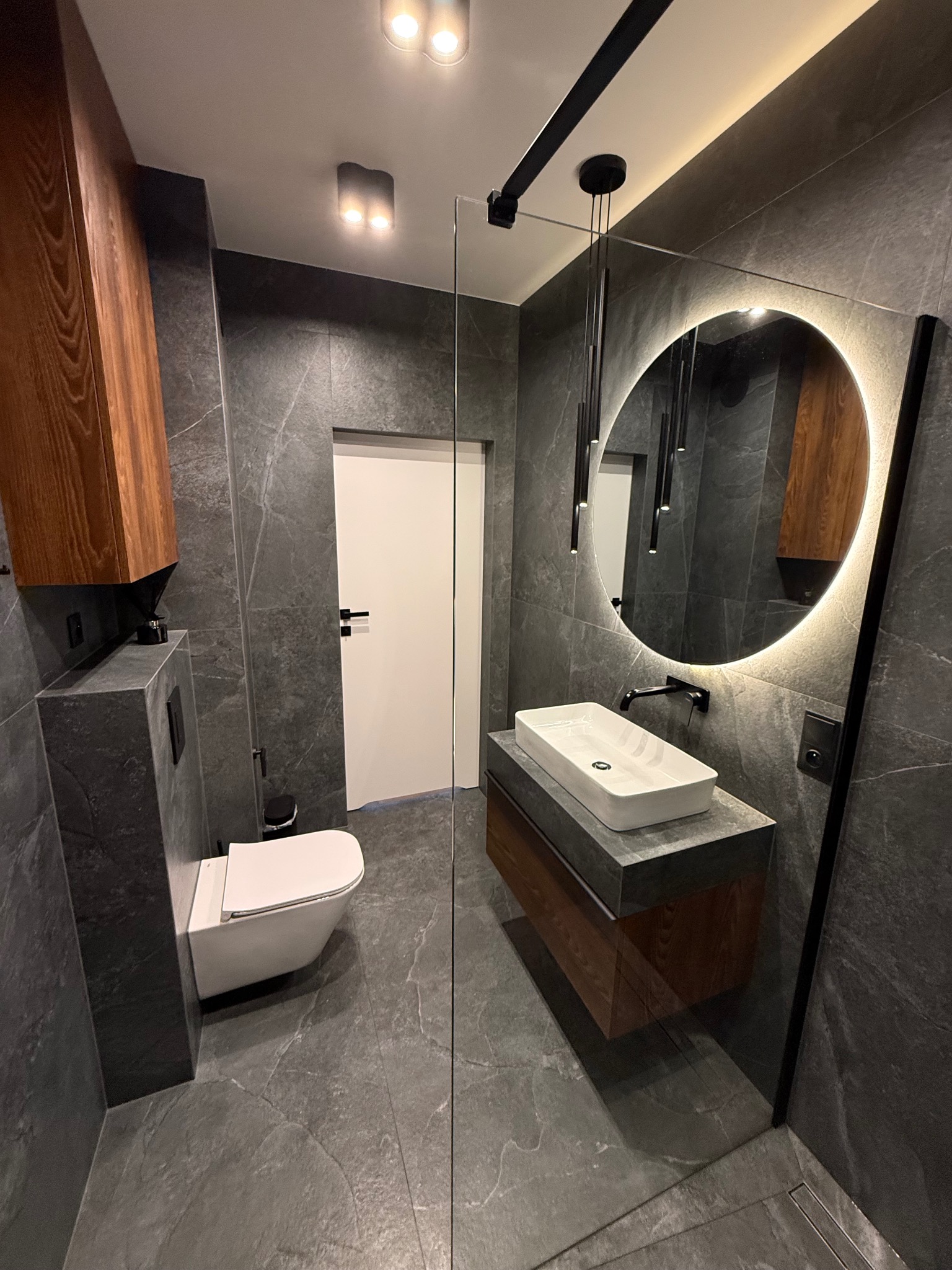 Nowoczesna łazienka z szarą płytką imitującą kamień, podświetlanym okrągłym lustrem i wiszącą toaletą. Minimalistyczny design z drewnianymi akcentami i szklaną kabiną.