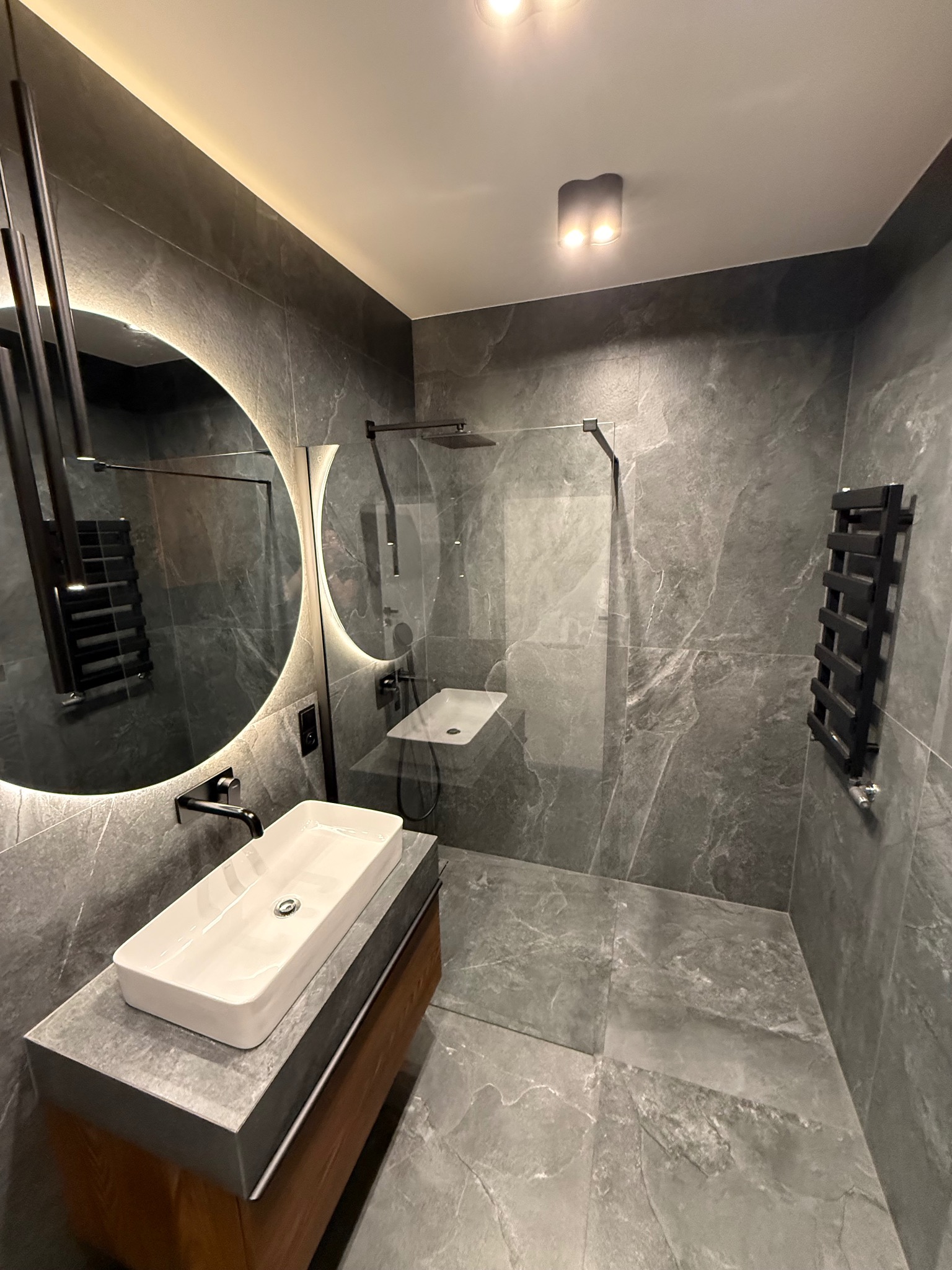 Nowoczesna łazienka w odcieniach szarości z okrągłym lustrem z podświetleniem LED, kabiną prysznicową typu walk-in i drewnianą szafką pod umywalką.