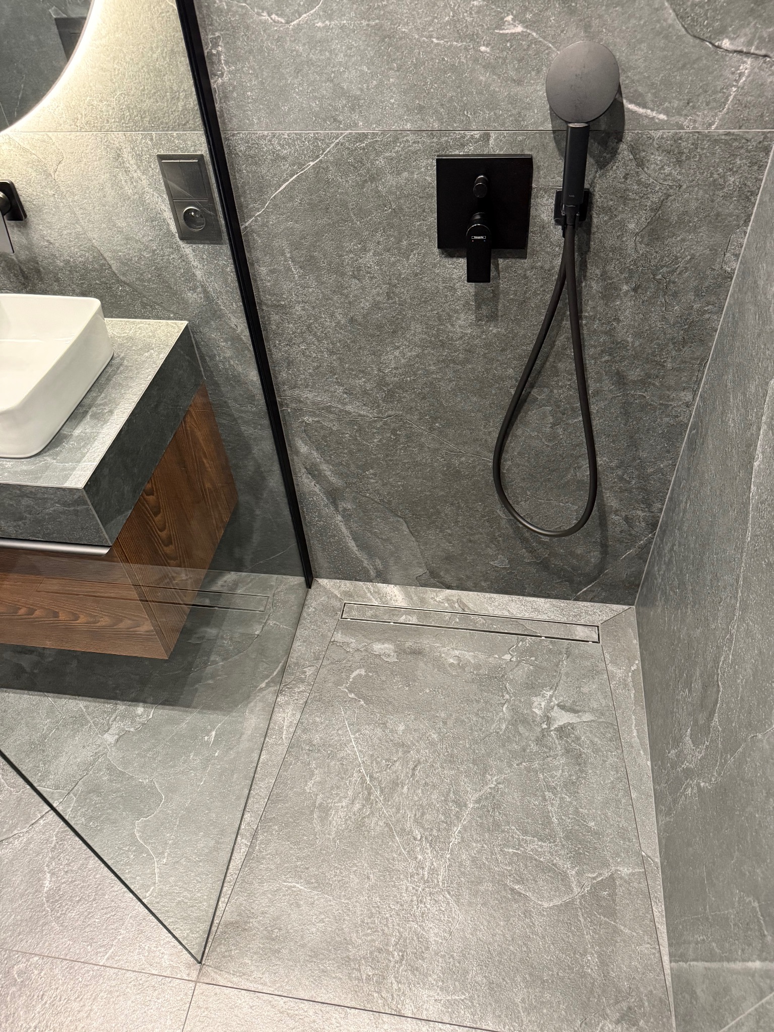 Nowoczesna łazienka z szarą płytką imitującą kamień, czarną armaturą i odpływem liniowym. Widoczna umywalka z drewnianą szafką i oświetlenie LED.