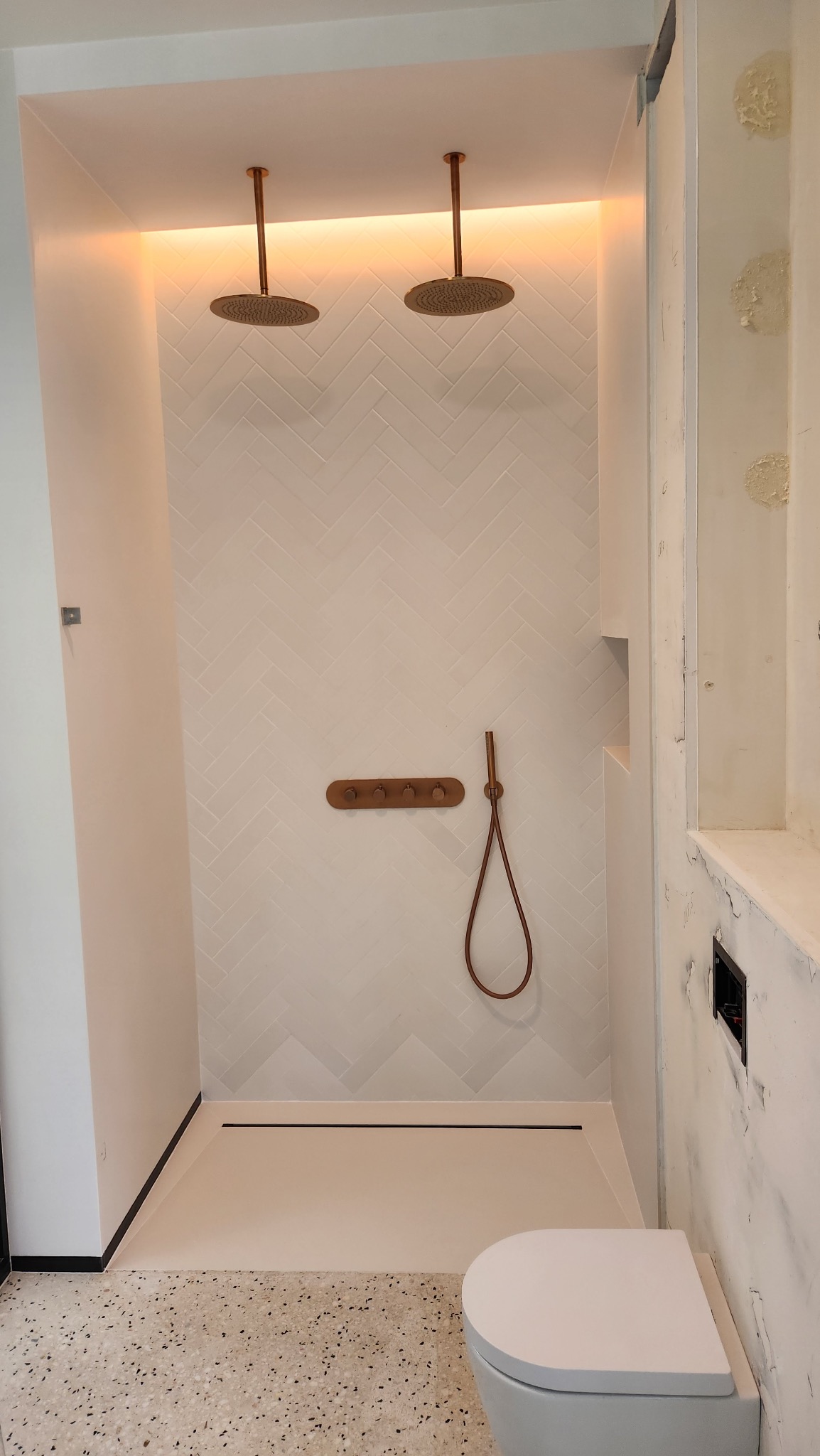 Nowoczesna łazienka z dwoma deszczownicami, białymi płytkami ułożonymi w jodełkę i elementami armatury w kolorze miedzi. Widoczna toaleta i odpływ liniowy.