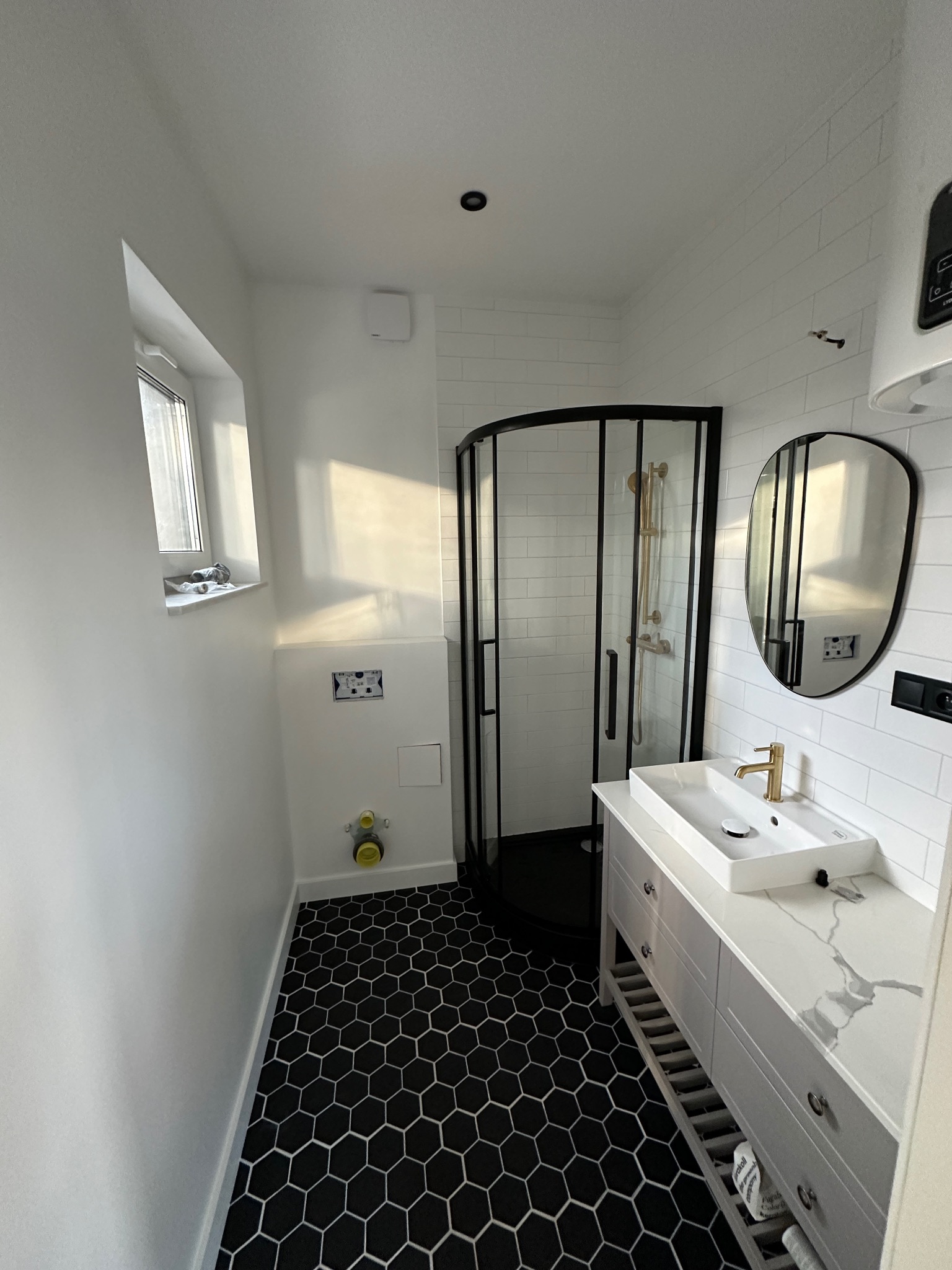 Nowoczesna łazienka z czarno-białą mozaiką na podłodze, kabiną prysznicową z czarnymi ramami i białą umywalką z baterią w kolorze złota.