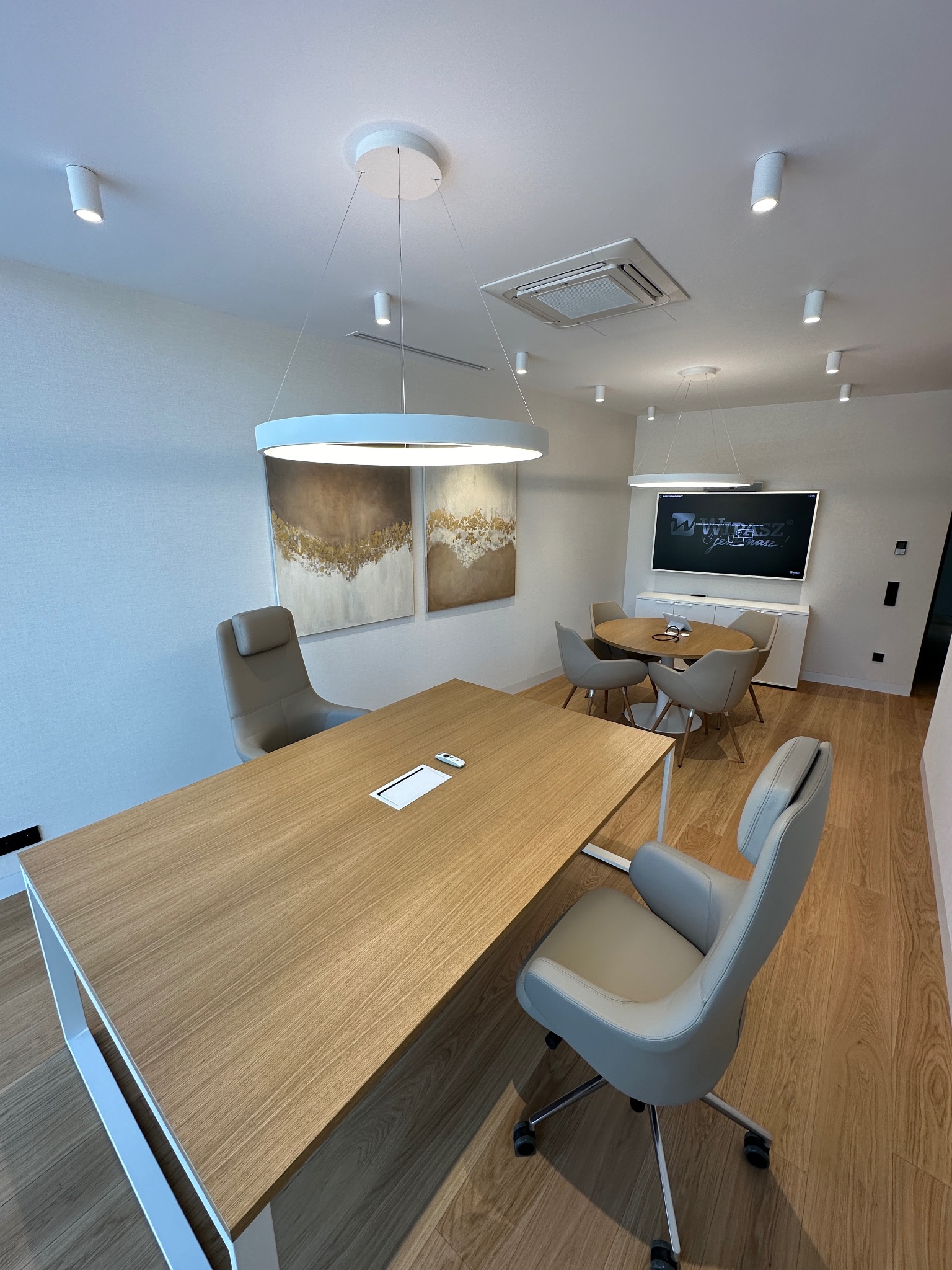 Nowoczesne biuro z drewnianym stołem, szarymi krzesłami i okrągłą lampą. Na ścianie wiszą obrazy, a w tle widać telewizor. Jasne wnętrze, minimalistyczny design.