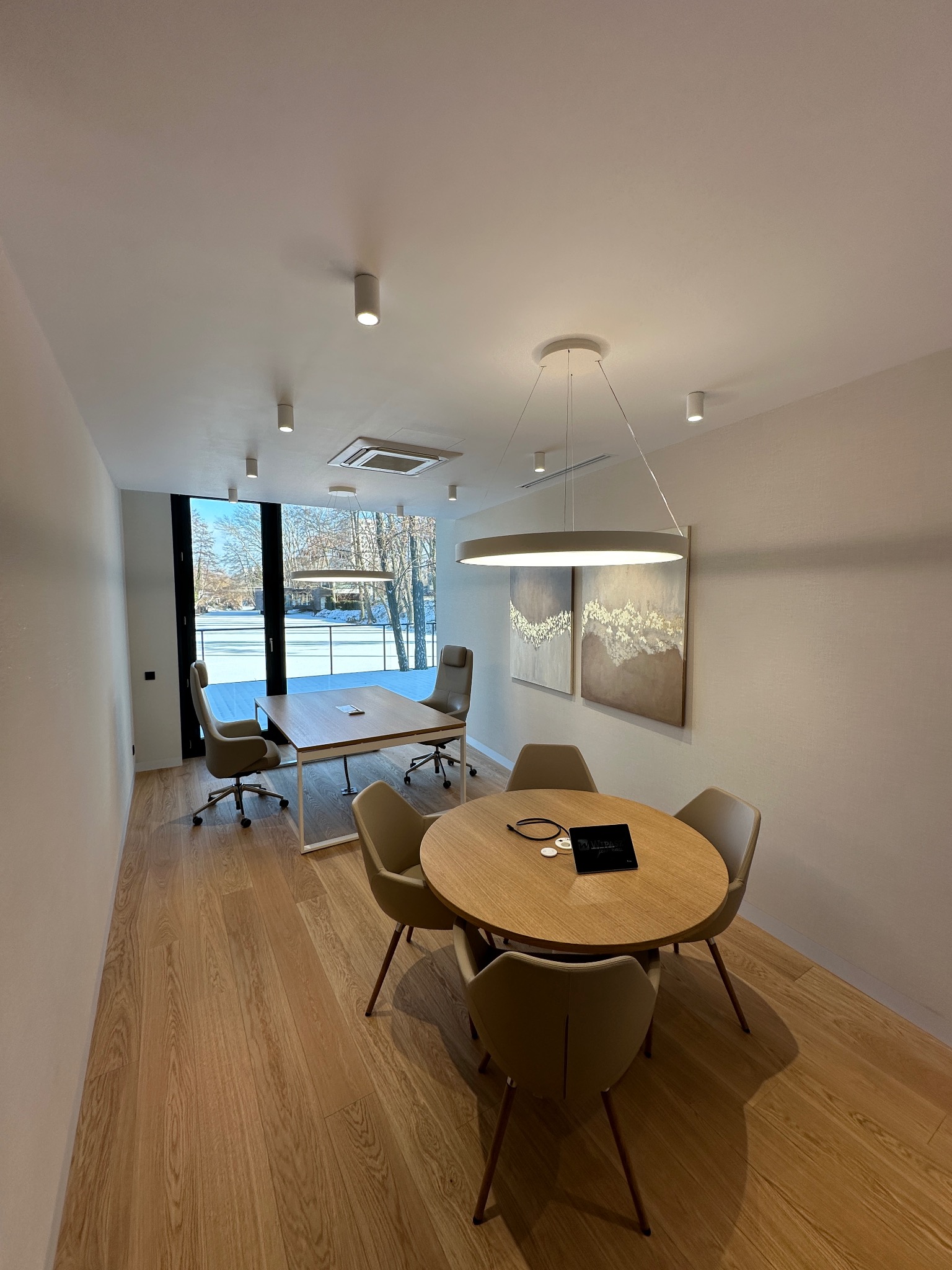 Nowoczesne biuro w Warszawie: drewniana podłoga, okrągły stół z krzesłami i widok na zimowy krajobraz za oknem. Minimalistyczne oświetlenie i obrazy.