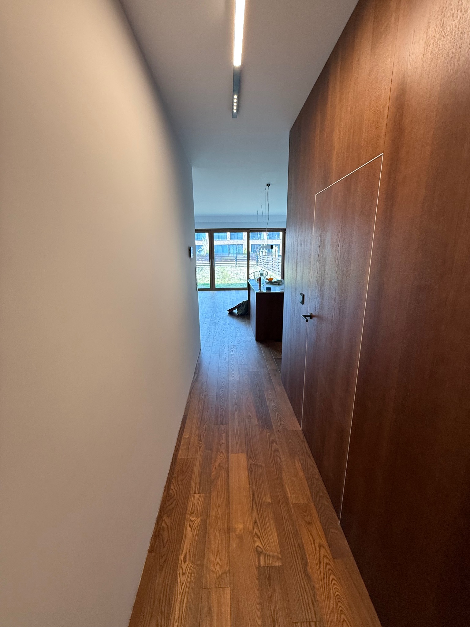 Długi korytarz z drewnianą podłogą i ścianą w odcieniach brązu. Ukryte drzwi w ścianie. W tle widoczne okno i wnętrze jasnego pomieszczenia.