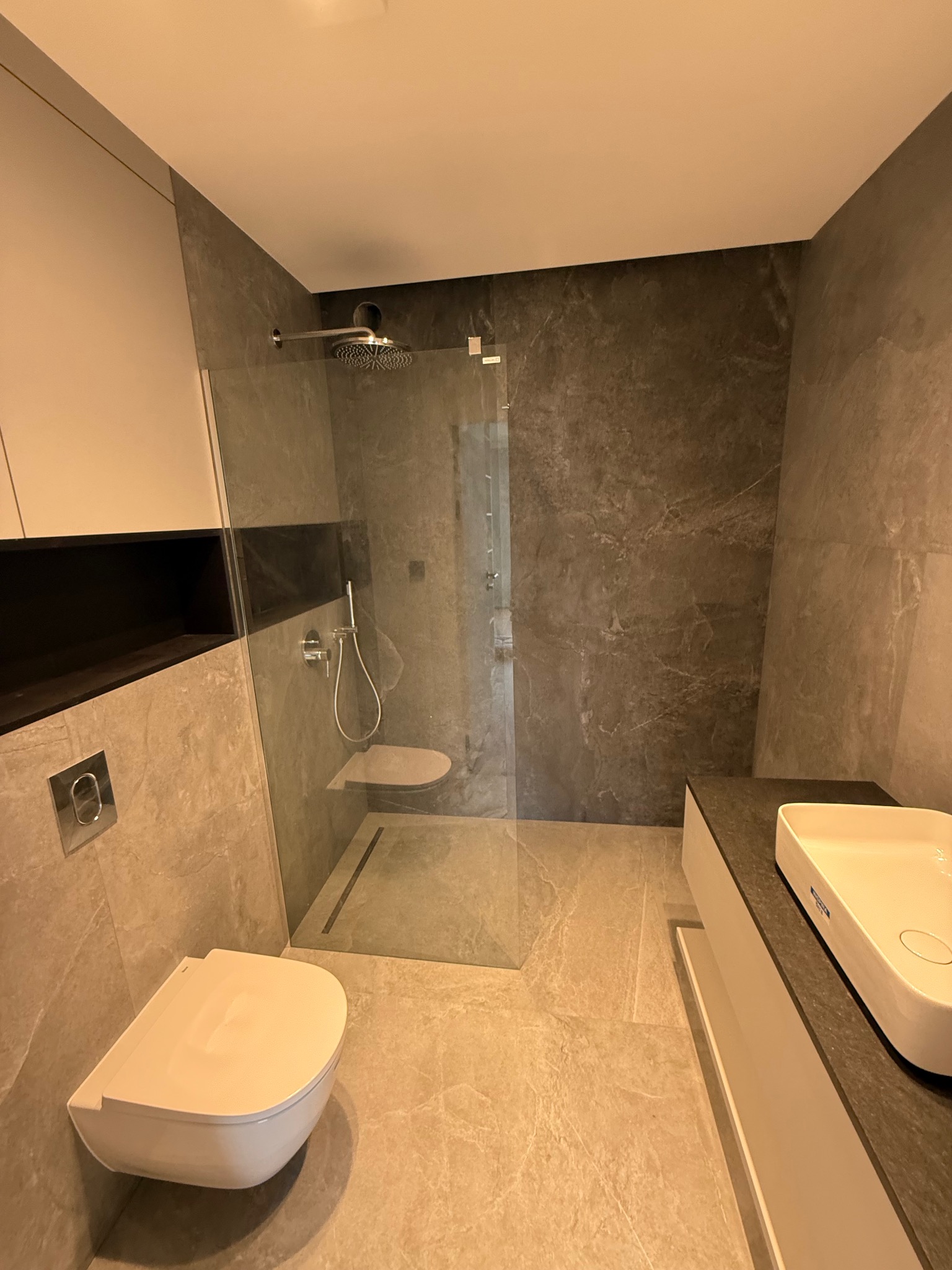 Nowoczesna łazienka z kabiną prysznicową typu walk-in, podwieszaną toaletą i umywalką nablatową. Szare płytki imitujące kamień na ścianach i podłodze.