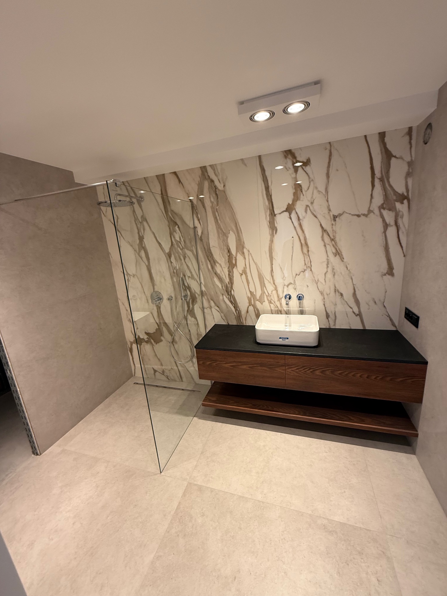 Nowoczesna łazienka z kabiną prysznicową typu walk-in, marmurową ścianą i drewnianą szafką pod umywalką. Minimalistyczny design i jasna kolorystyka.