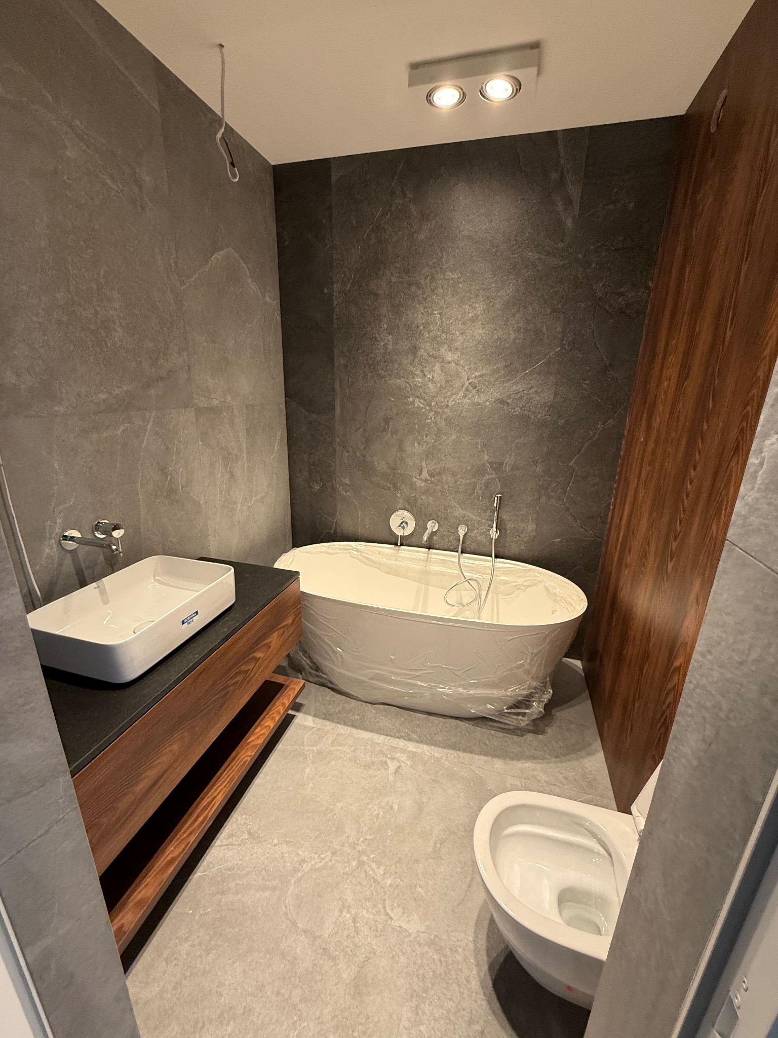 Nowoczesna łazienka w szarościach z drewnianymi akcentami. Wanna owinięta folią ochronną, umywalka na szafce z drewnianymi półkami i podwieszana toaleta.