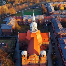 Jan Pasterny - Panorama z lotu ptaka na ceglany kościół z pomarańczowym dachem i białymi wieżami, otoczony budynkami mieszkalnymi i zielenią w jesiennym słońcu.