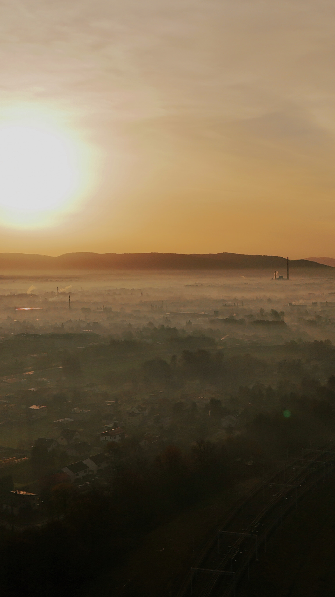 Panorama miasta spowitego poranną mgłą, z widocznym wschodzącym słońcem i górami w tle, uchwycona z góry.