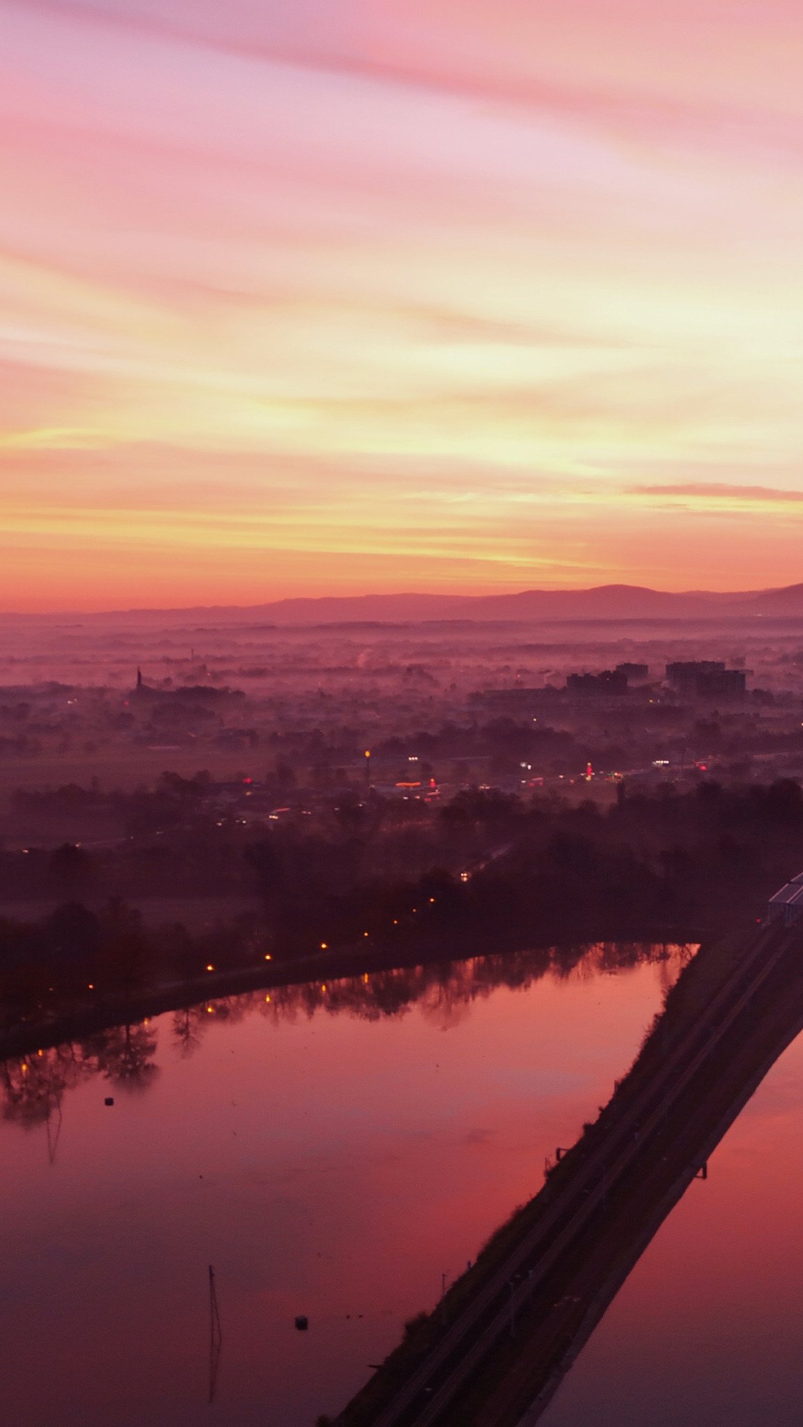 Panoramiczny widok na miasto w porannej mgle, z rzeką odbijającą różowo-pomarańczowe niebo o wschodzie słońca, widoczne budynki i wieża kościoła w oddali.