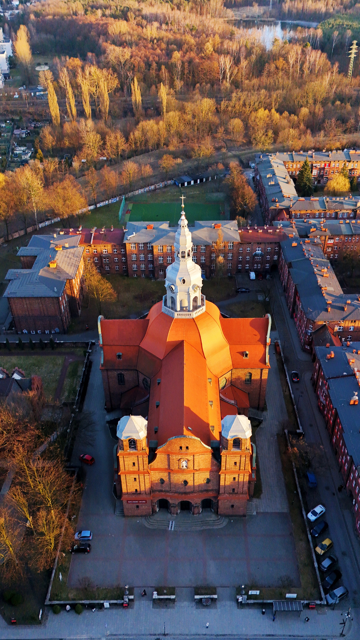 Panorama z lotu ptaka na ceglany kościół z pomarańczowym dachem i białymi wieżami, otoczony budynkami mieszkalnymi i zielenią w jesiennym słońcu.