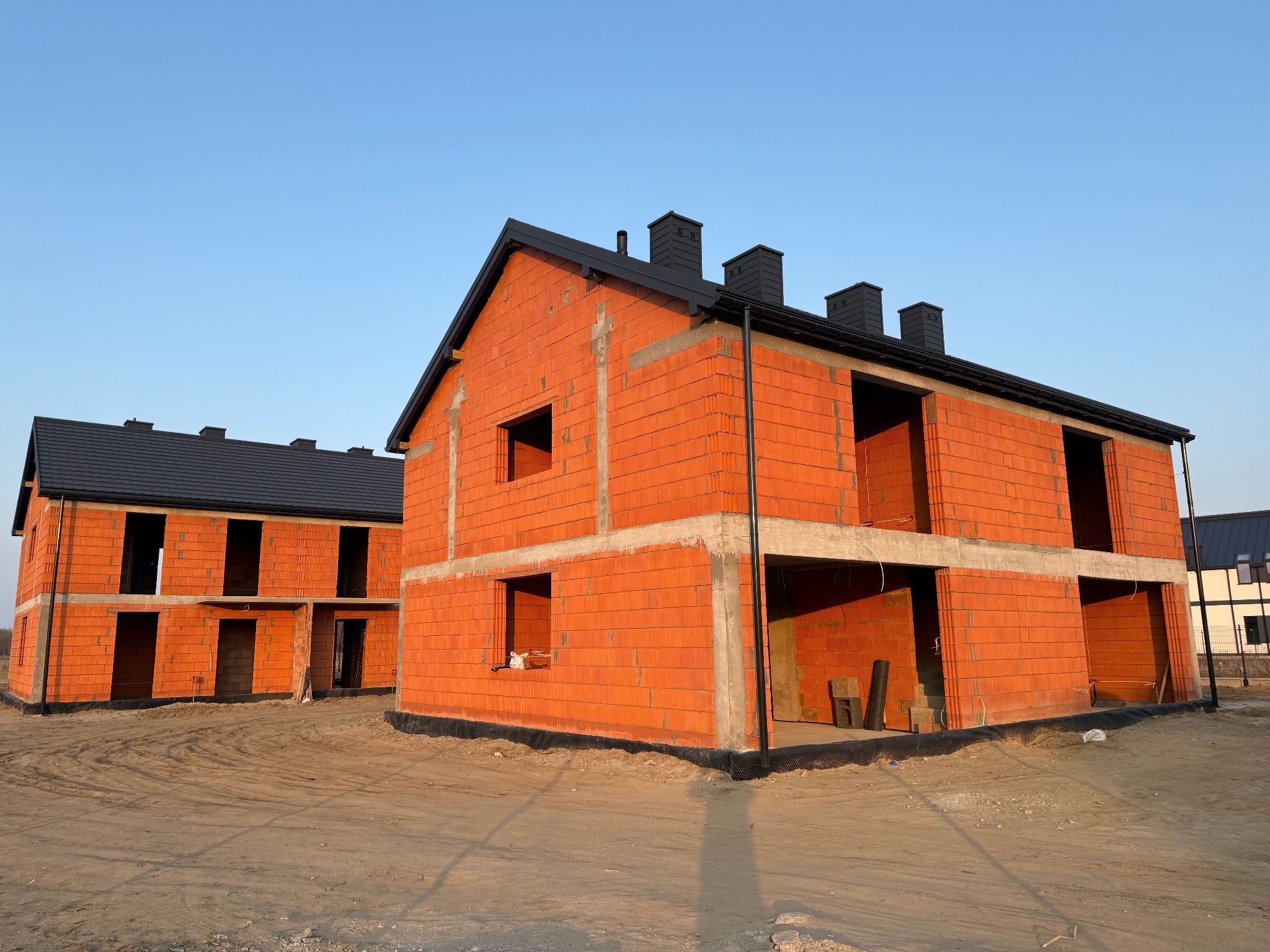 Surowy stan bliźniaczych domów z czerwonej cegły z czarnymi dachami i kominami, widoczne z zewnątrz w słoneczny dzień, teren wokół przygotowany pod dalsze prace budowlane.