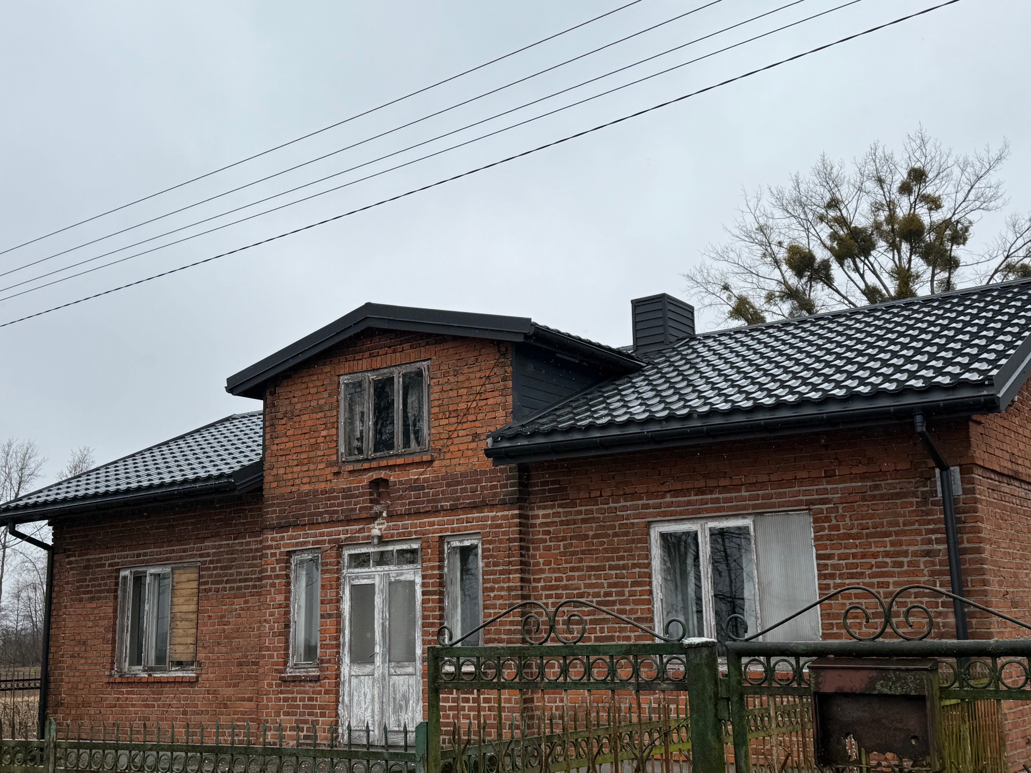 Ceglany dom z nowym, czarnym dachem z dachówki ceramicznej, widoczne stare okna i brama, pochmurne niebo.