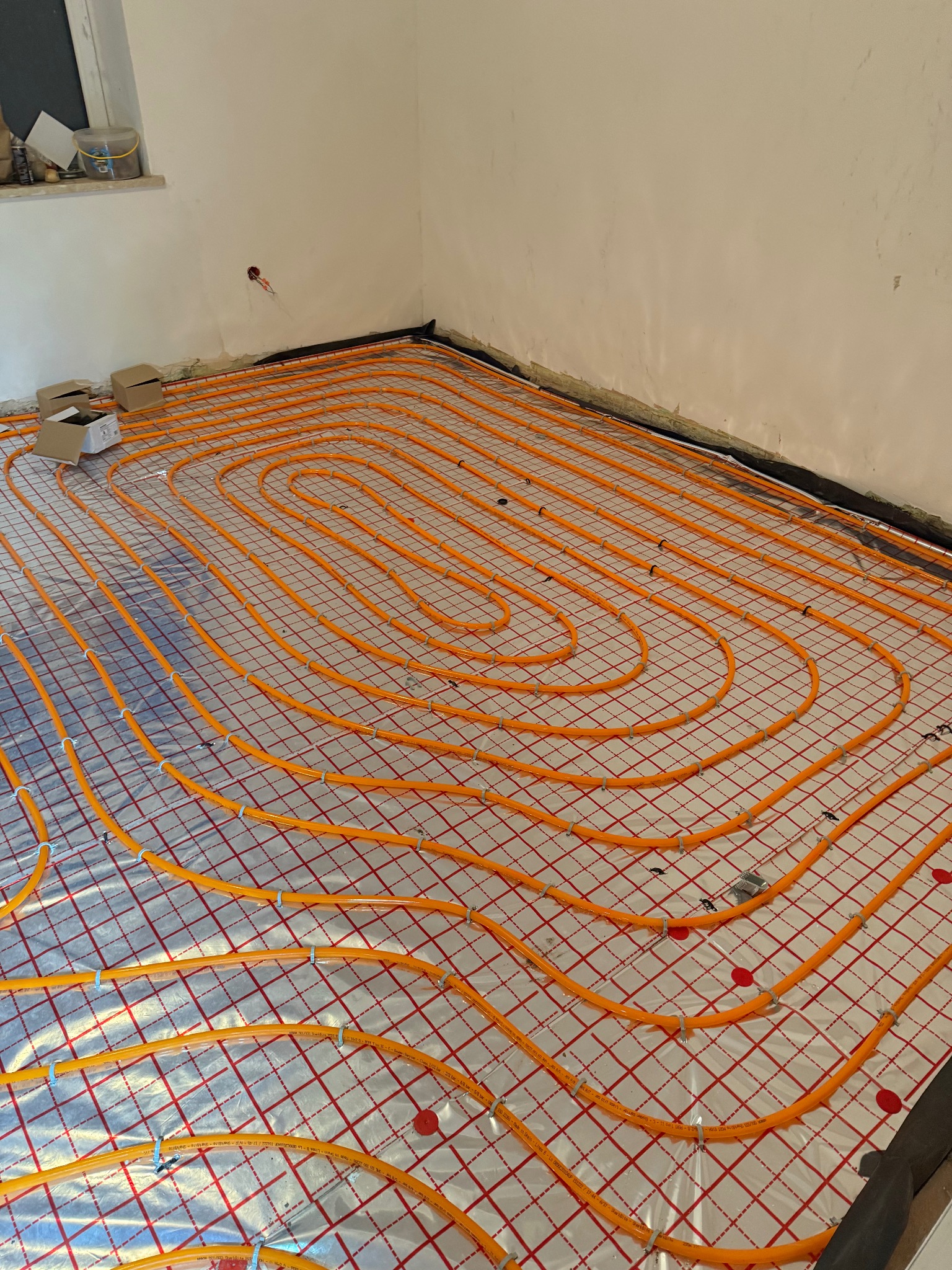 Pomarańczowe rury ogrzewania podłogowego ułożone spiralnie na izolacji z siatką w nowym budynku, widoczne kartony i elementy instalacji przy ścianie.