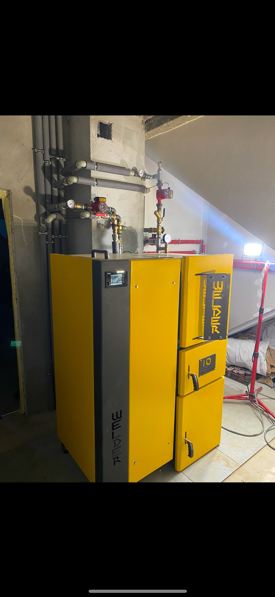 Żółto-szary piec CO marki Weljak z podłączoną instalacją hydrauliczną z rur miedzianych i plastikowych w pomieszczeniu z surowym betonem.