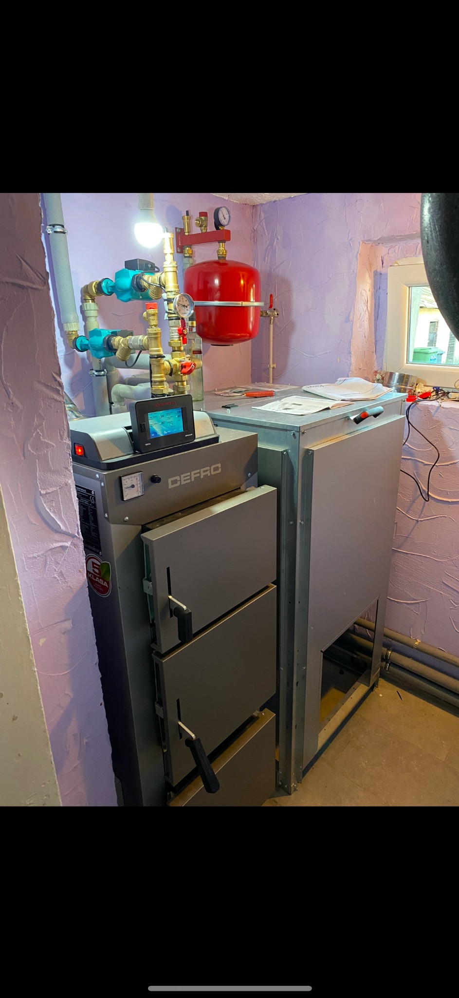 Szary piec CO marki DEFRO z podłączoną instalacją hydrauliczną i czerwonym zbiornikiem wyrównawczym w pomieszczeniu z fioletowymi ścianami o nierównej fakturze.