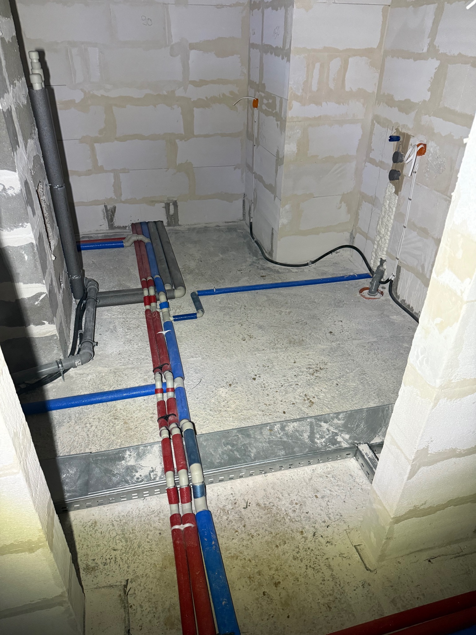 Surowe wnętrze w trakcie prac hydraulicznych: widoczne rury instalacyjne w kolorach czerwonym i niebieskim, prowadzone wzdłuż surowych ścian z betonu komórkowego, oraz elementy instalacji grzewczej...