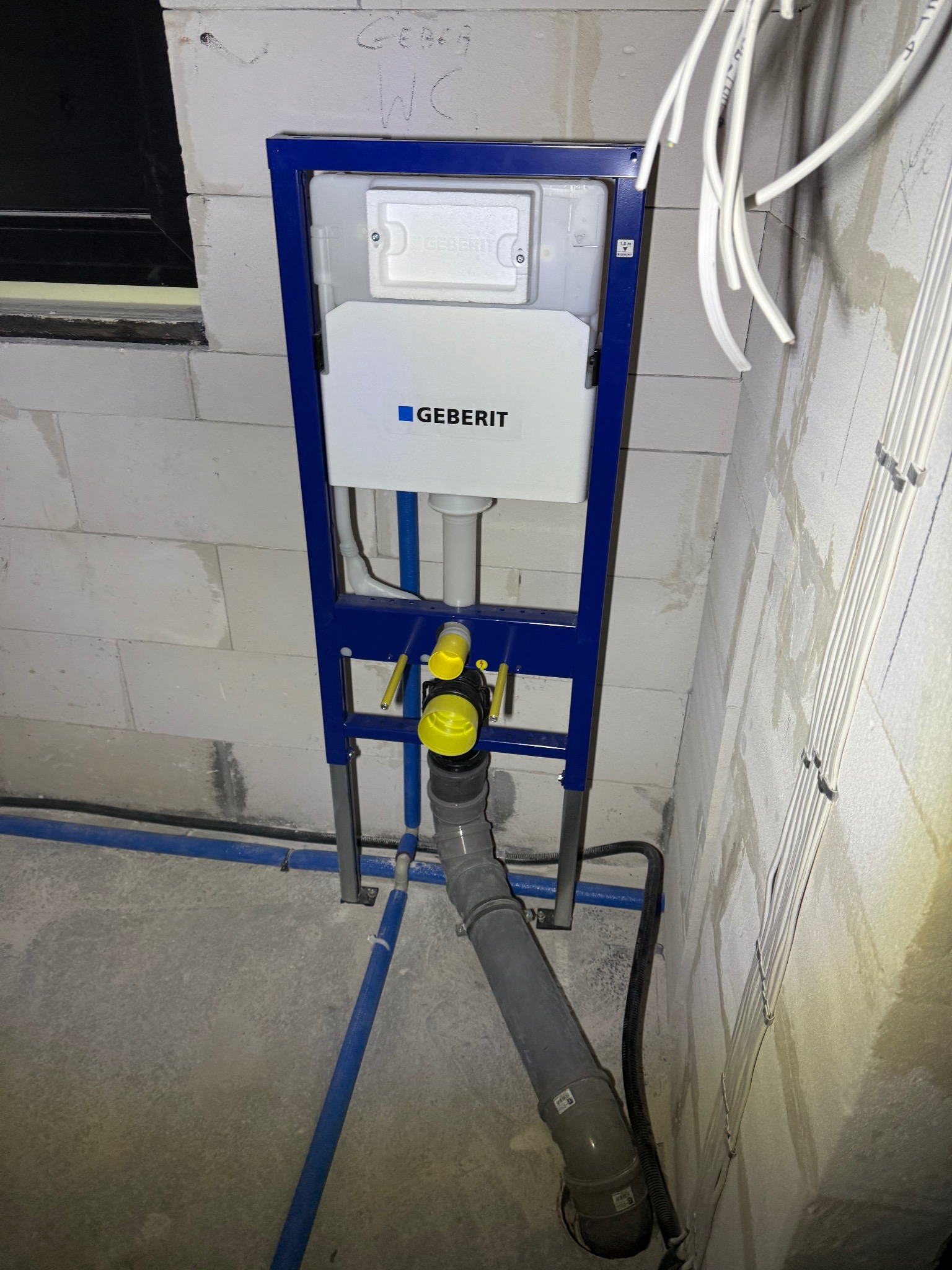 Stelaż podtynkowy Geberit zamontowany przy ścianie z bloczków, widoczne niebieskie rury, szare rury kanalizacyjne i kable elektryczne w listwach.