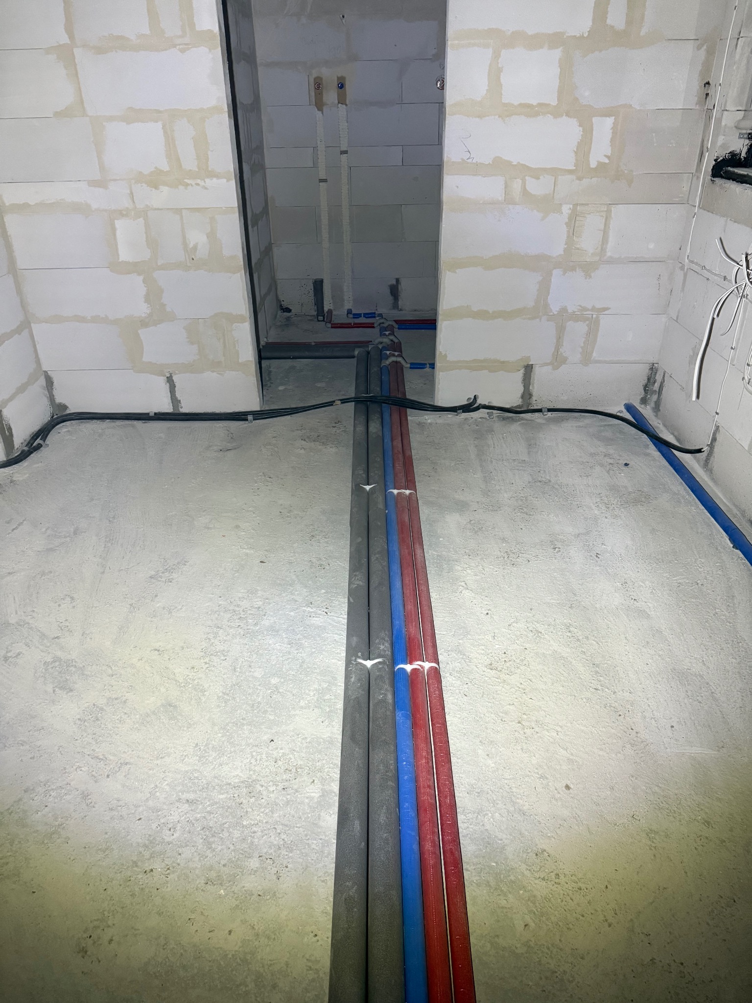 Surowy stan pomieszczenia w trakcie prac instalacyjnych: rury grzewcze i wodne w izolacji prowadzone wzdłuż ściany z bloczków, widoczne kable elektryczne i przygotowane podejścia do armatury.