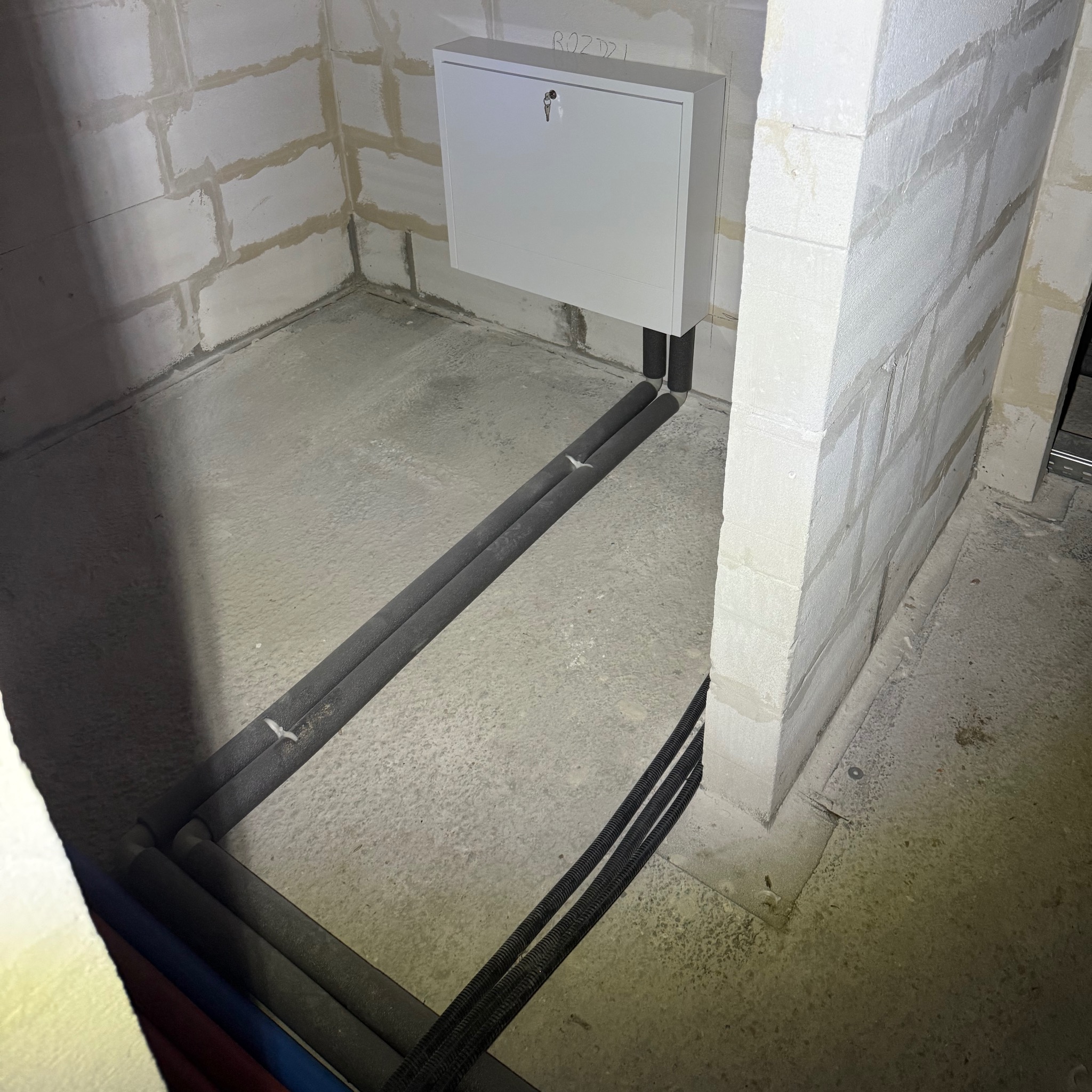 Instalacja ogrzewania podłogowego w trakcie realizacji w Bodzanowie: widoczne rury grzewcze z izolacją termiczną ułożone na surowym betonie w pomieszczeniu z bloczków betonowych, szafka rozdzielcza...